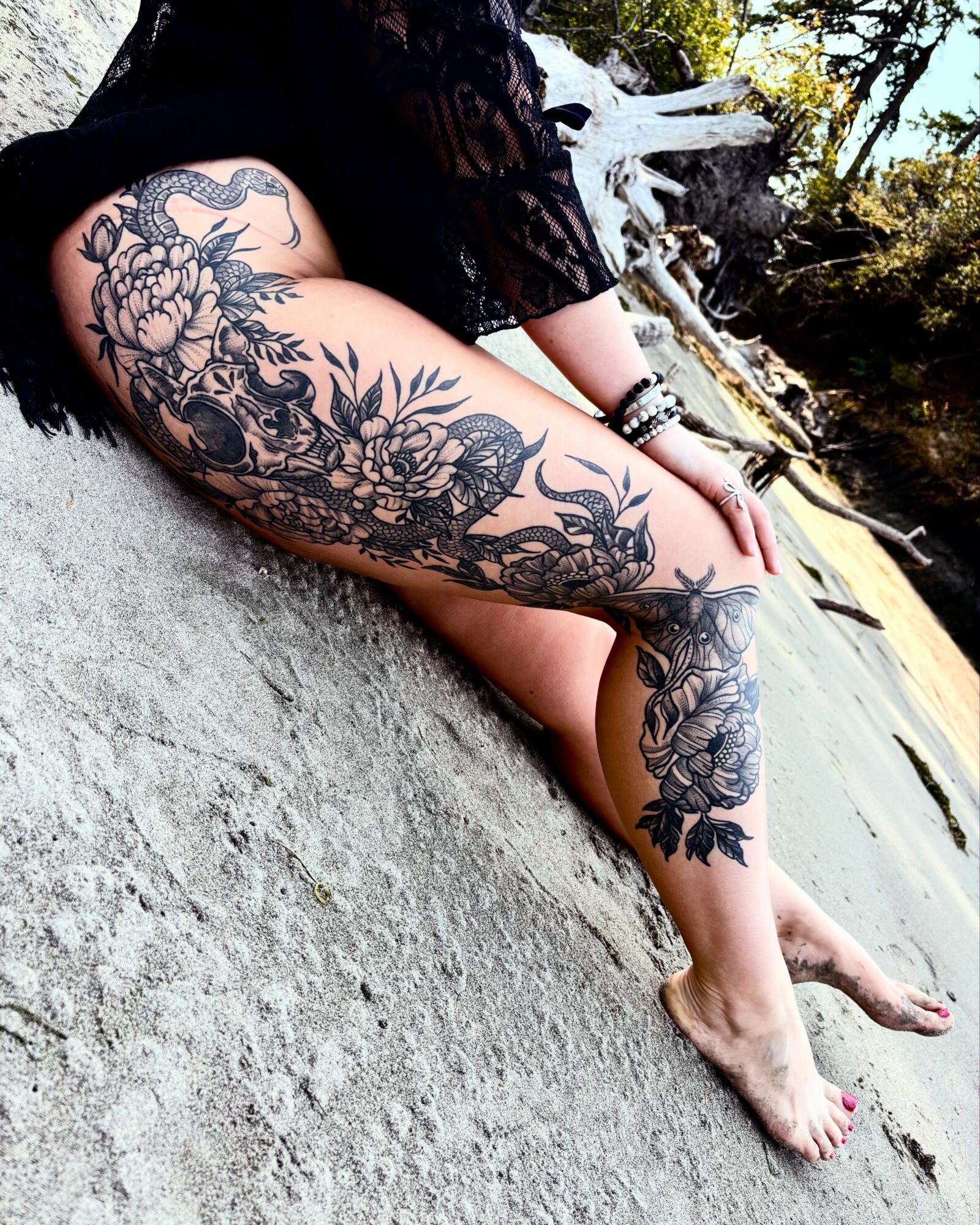 Person with a large floral tattoo on their arm, sitting on a rock near water, outdoors, with trees in the background.
