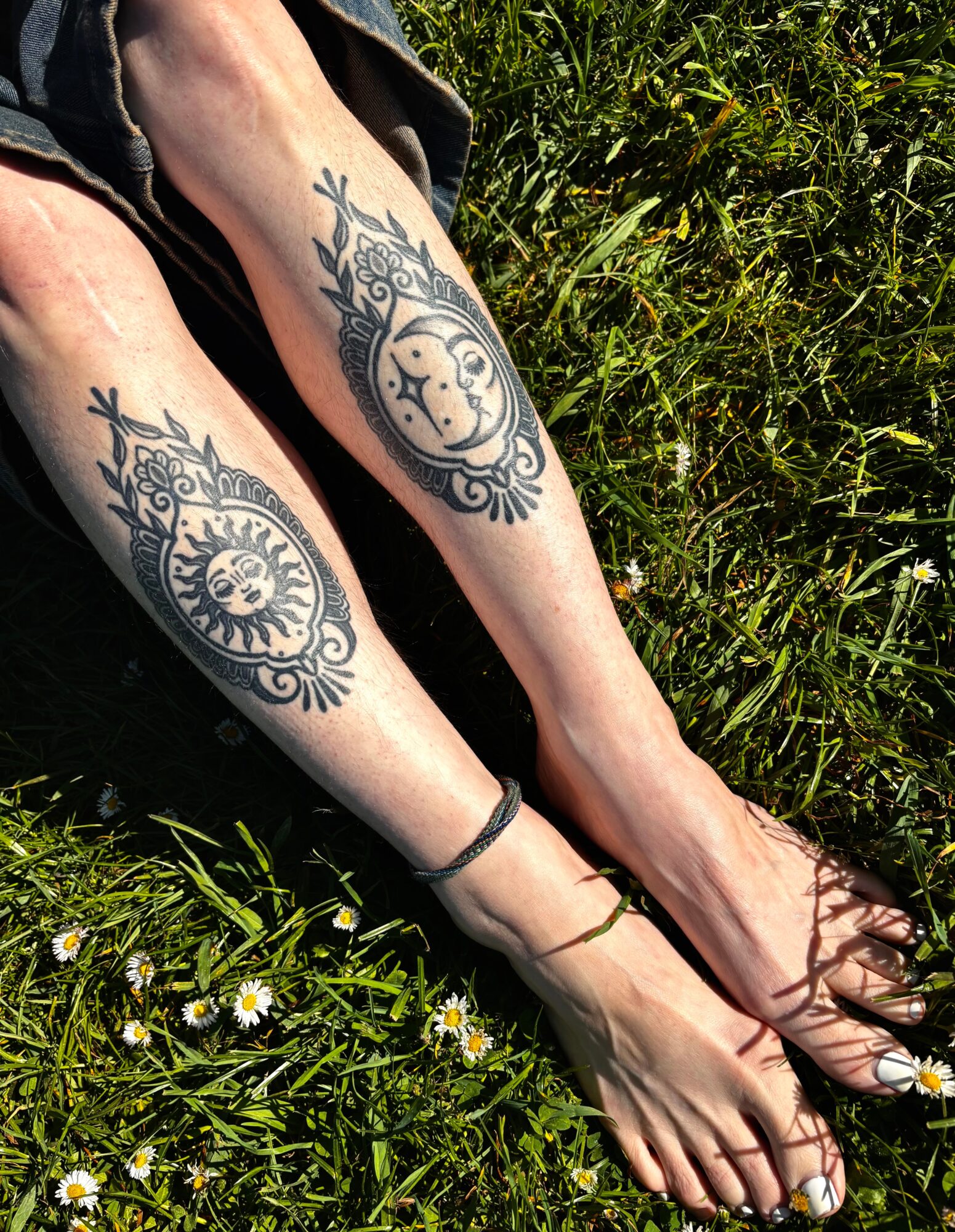 Two arms with sun and moon tattoos resting on grass, one with a bracelet, sunlight, and sandals visible.