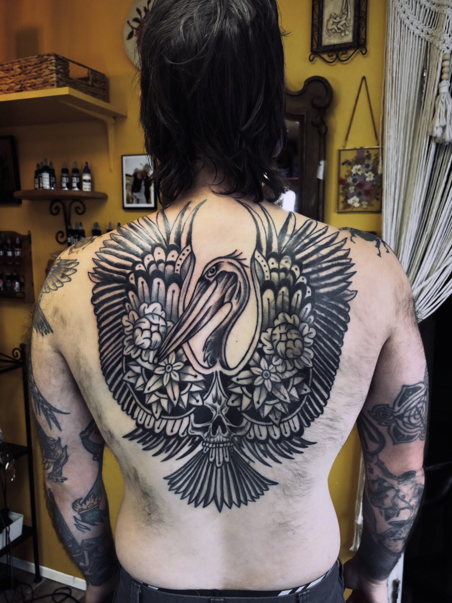 Back view of a person with a large swan tattoo surrounded by floral and feather designs, covering the upper back and shoulders.