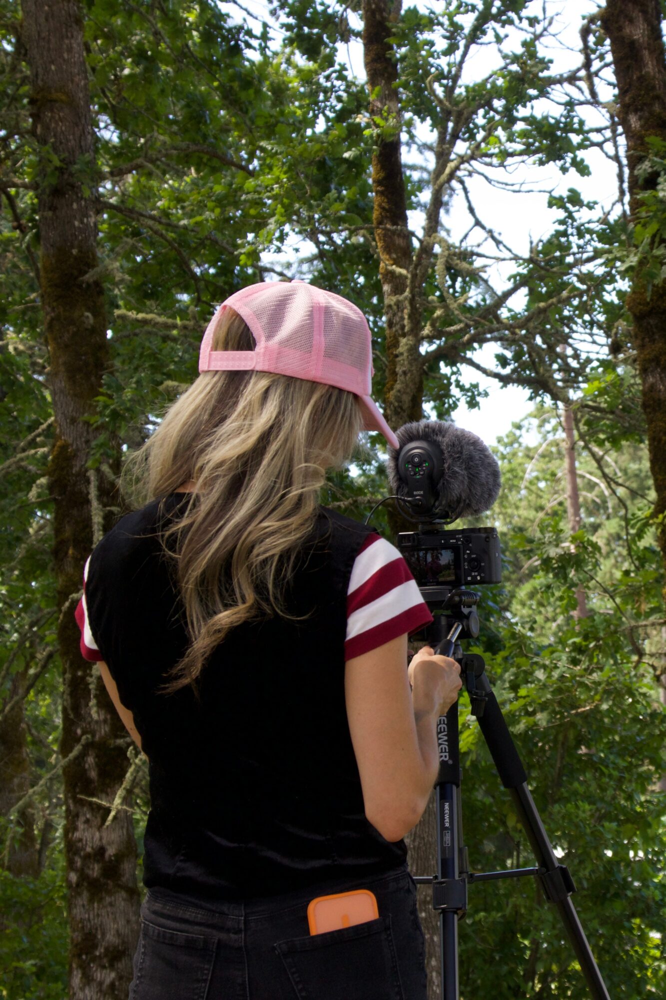 Person with long hair wearing a pink cap and black shirt with red and white striped sleeves, operating a camera outdoors.