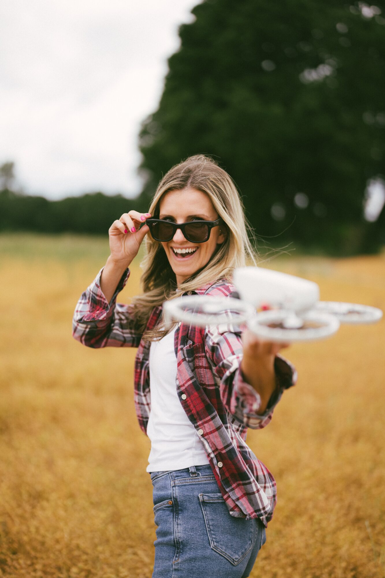 Woman smiling and adjusting sunglasses outdoors in a field with trees in the background.