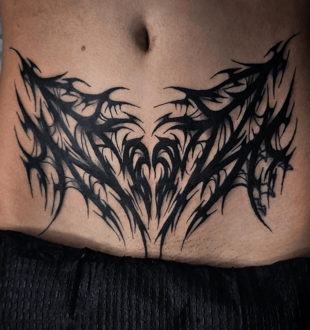Black tribal-style tattoo across the lower abdomen with sharp, jagged lines and symmetrical design.