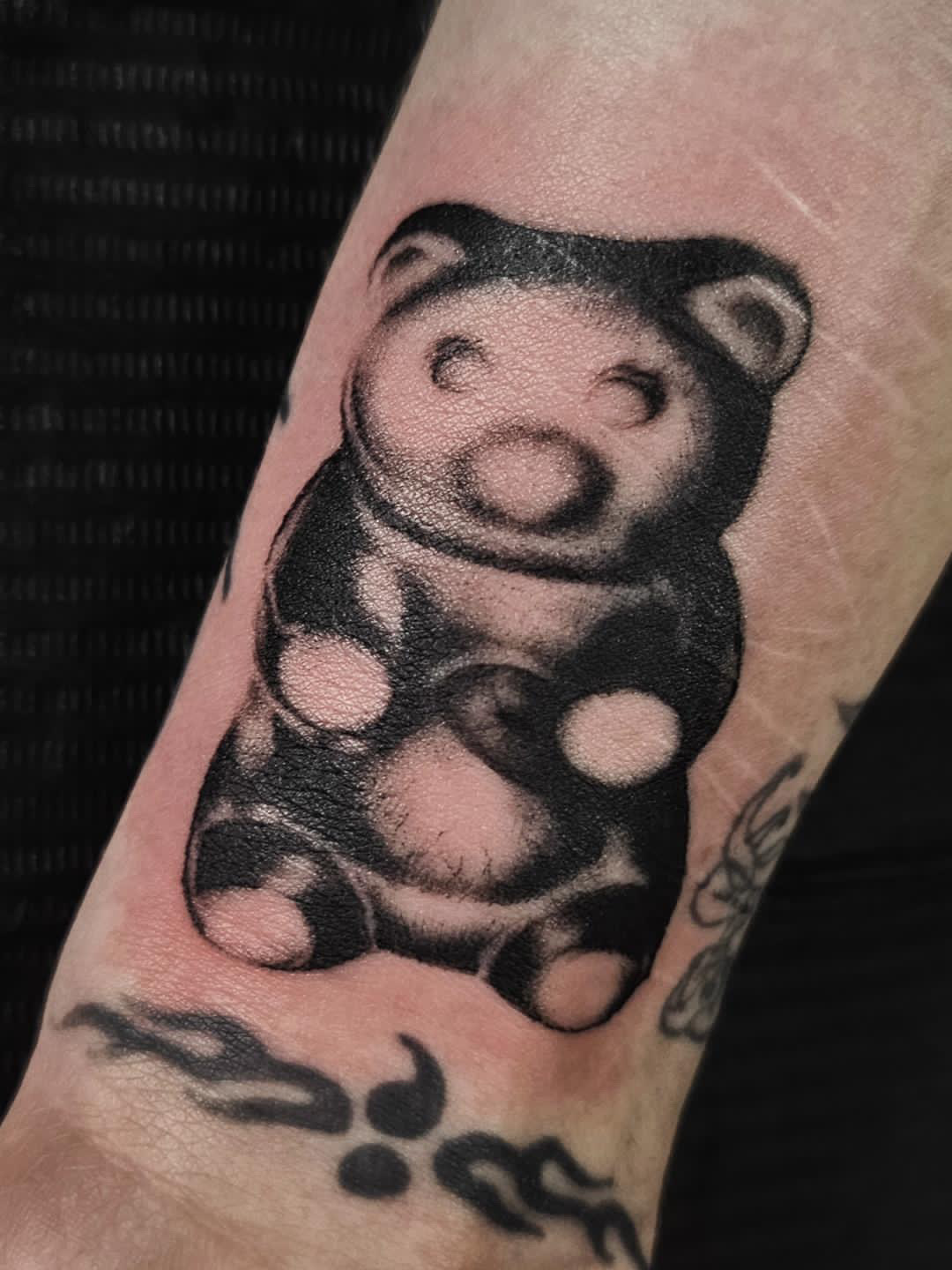 Black and gray tattoo of a cartoon-style bear with round ears and a smiling face on skin.