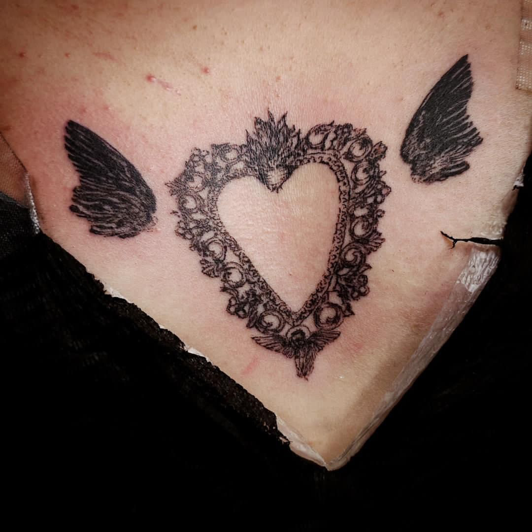 Tattoo of a heart-shaped lace design with two black wings on each side on skin.