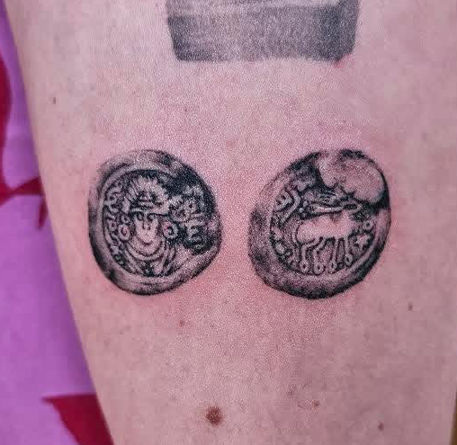 Tattoo of two ancient coin-like symbols, one with a face and the other with a deer, on skin.