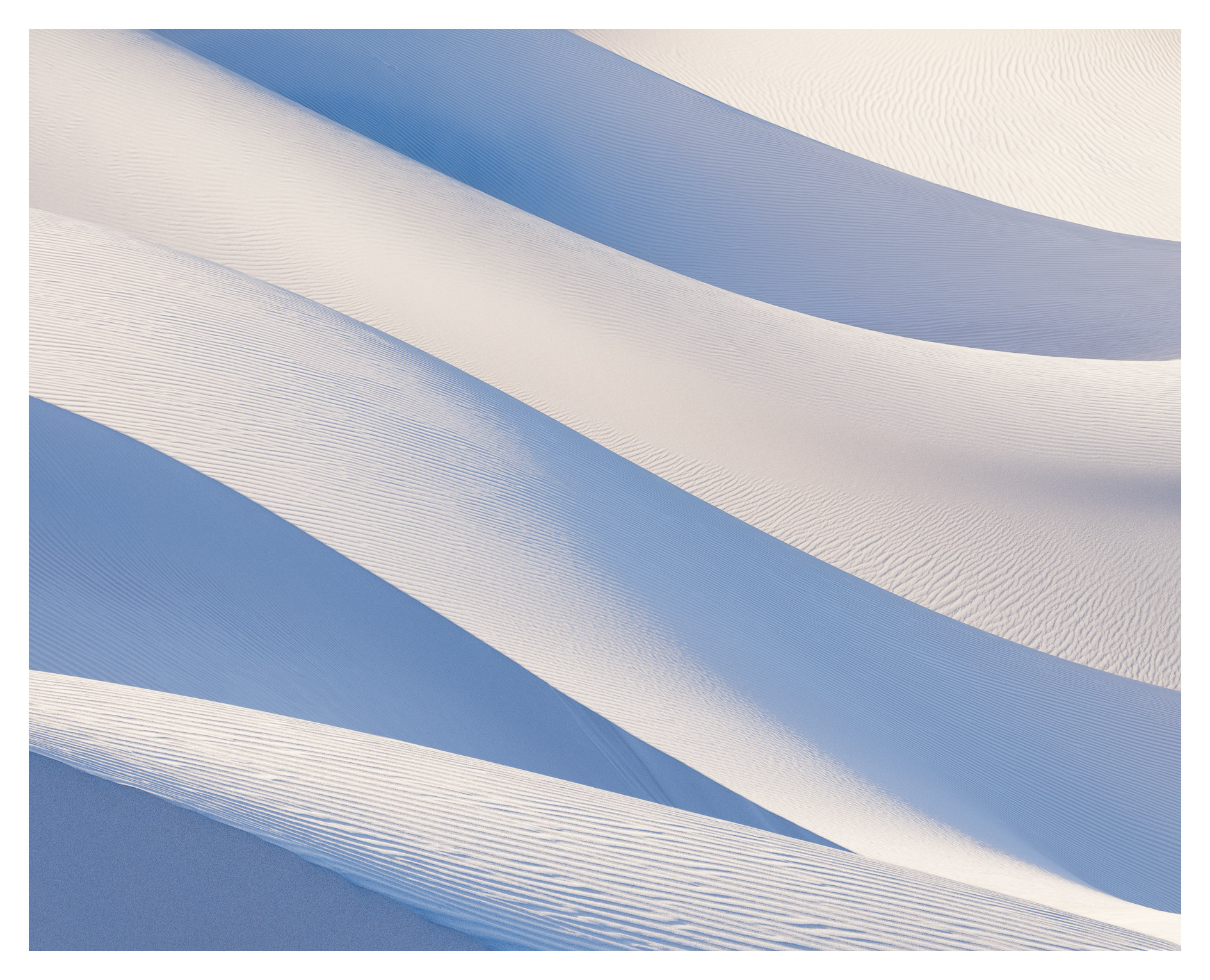 Sand dunes with smooth, curved shapes and shadows, in shades of white and blue, under a clear sky.
