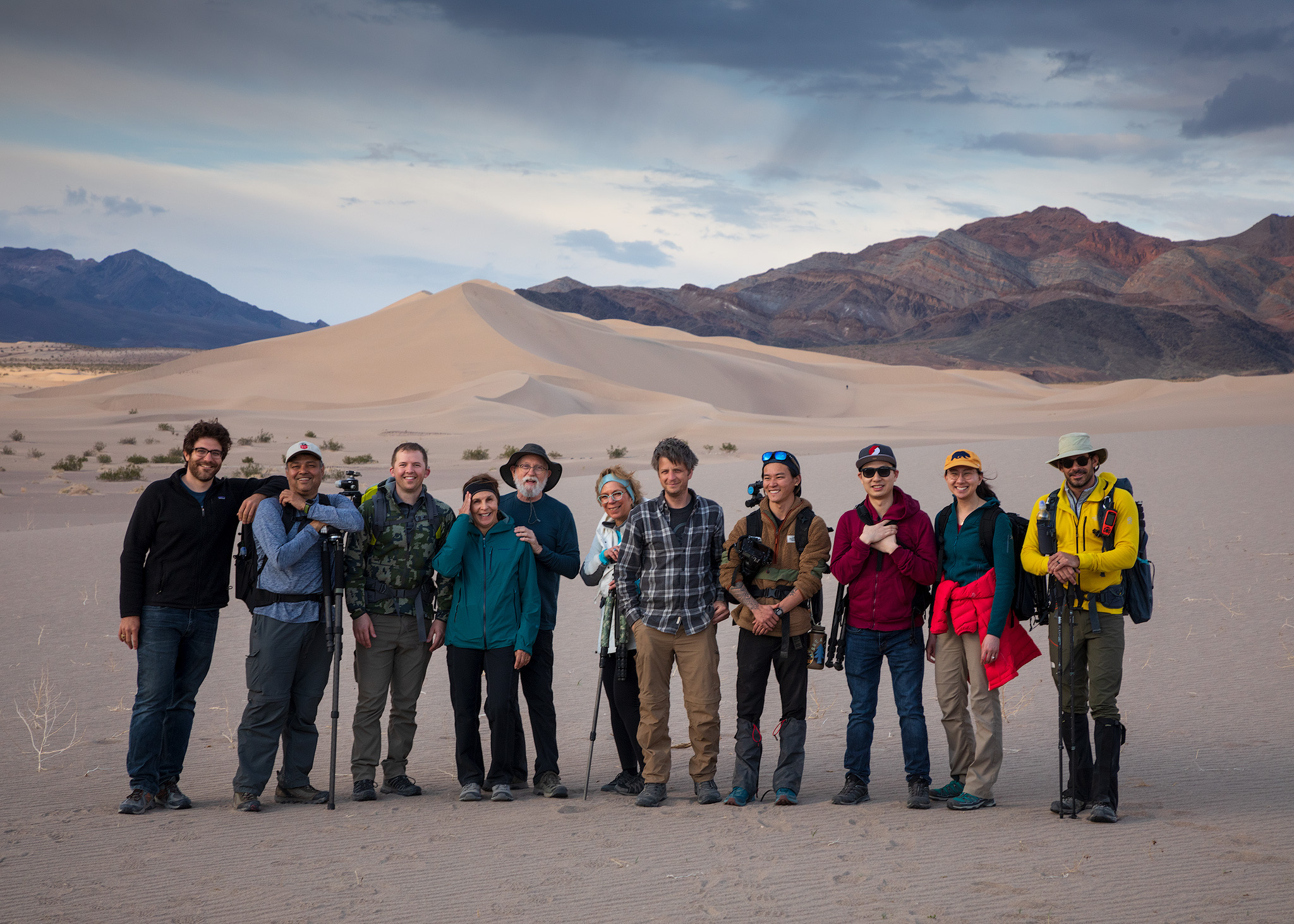 Group of ten people standing in desert with mountains and sand dunes in background, some holding hiking poles, wearing outdoor clothing.