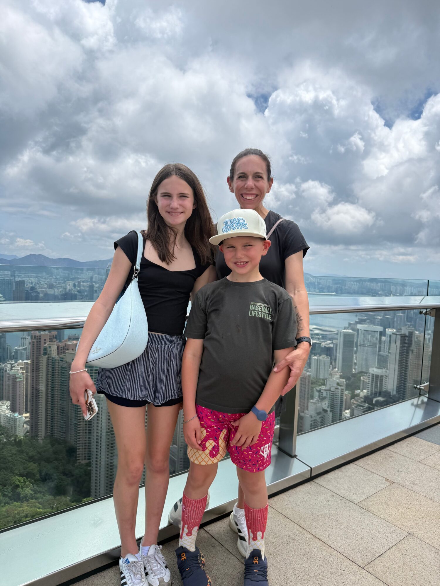 Three people standing on a balcony with a city skyline and cloudy sky in the background.