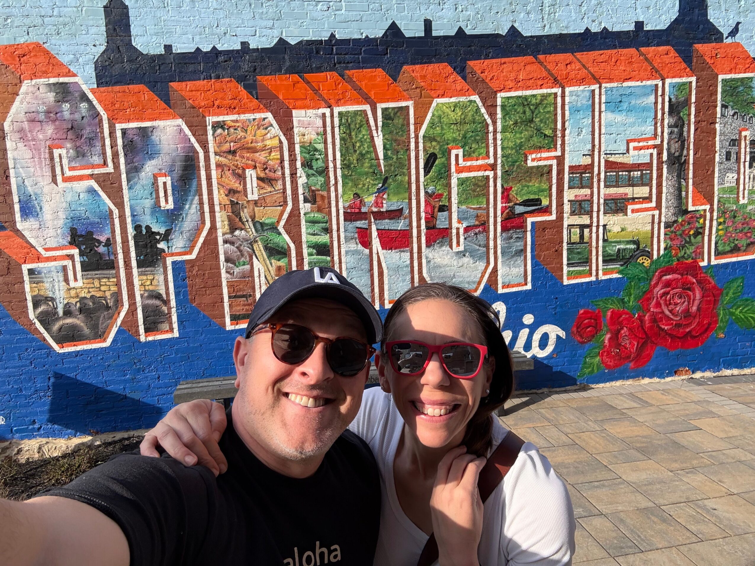 Two people smiling and taking a selfie in front of a colorful mural with large letters and a red rose.