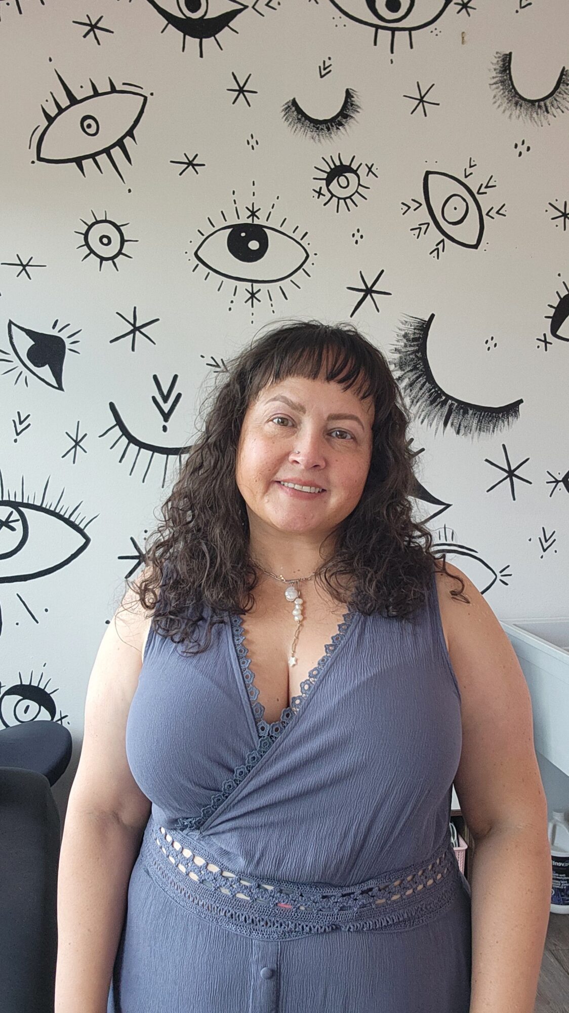 Woman with curly hair wearing a blue top, standing in front of a wall with eye and moon illustrations.