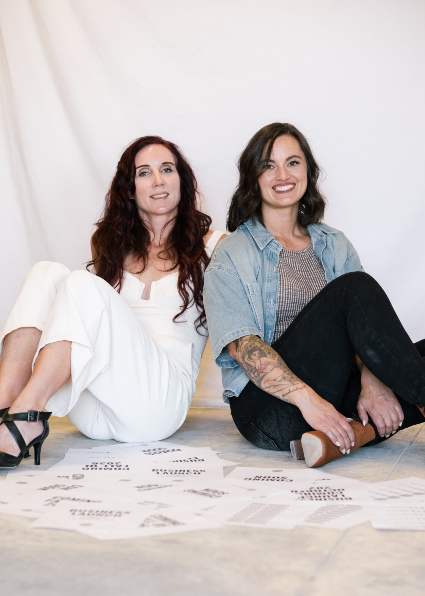 Two women sitting on the floor with papers scattered in front, smiling, against a plain white background.