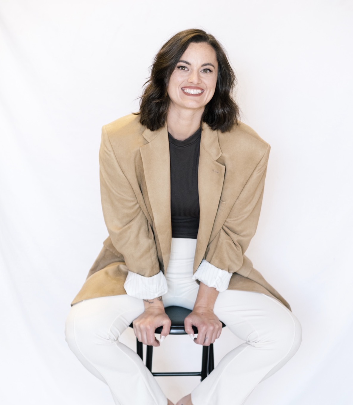 Woman with shoulder-length dark hair smiling, wearing a beige blazer, black top, and white pants, sitting on a stool against a white background.