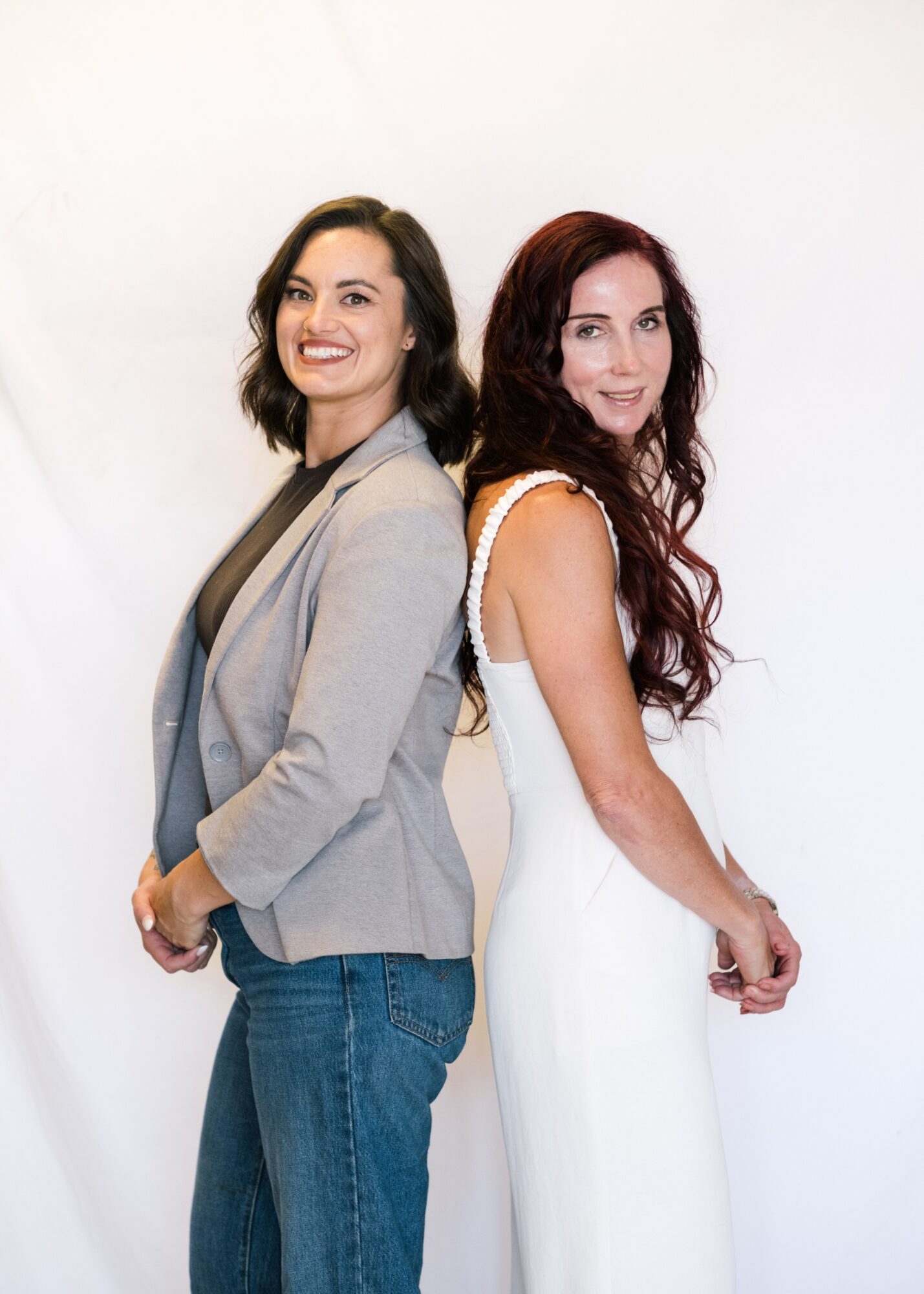 Two women stand back-to-back, smiling, against a plain white background. One wears a gray blazer and jeans, the other a white dress.