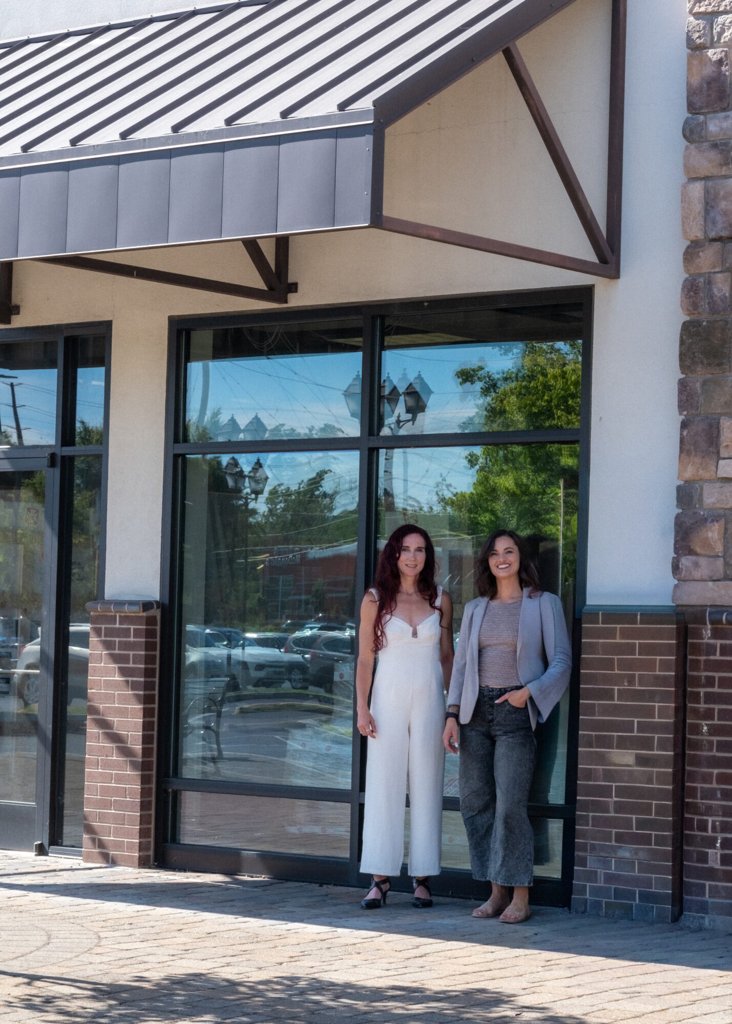 Two women stand outside a building with large glass windows, brick and stone exterior, and a metal awning, reflected in the glass.