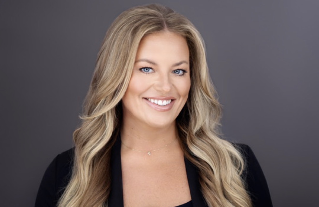 Portrait of a smiling woman with long wavy blonde hair, wearing a black top, against a gray background.