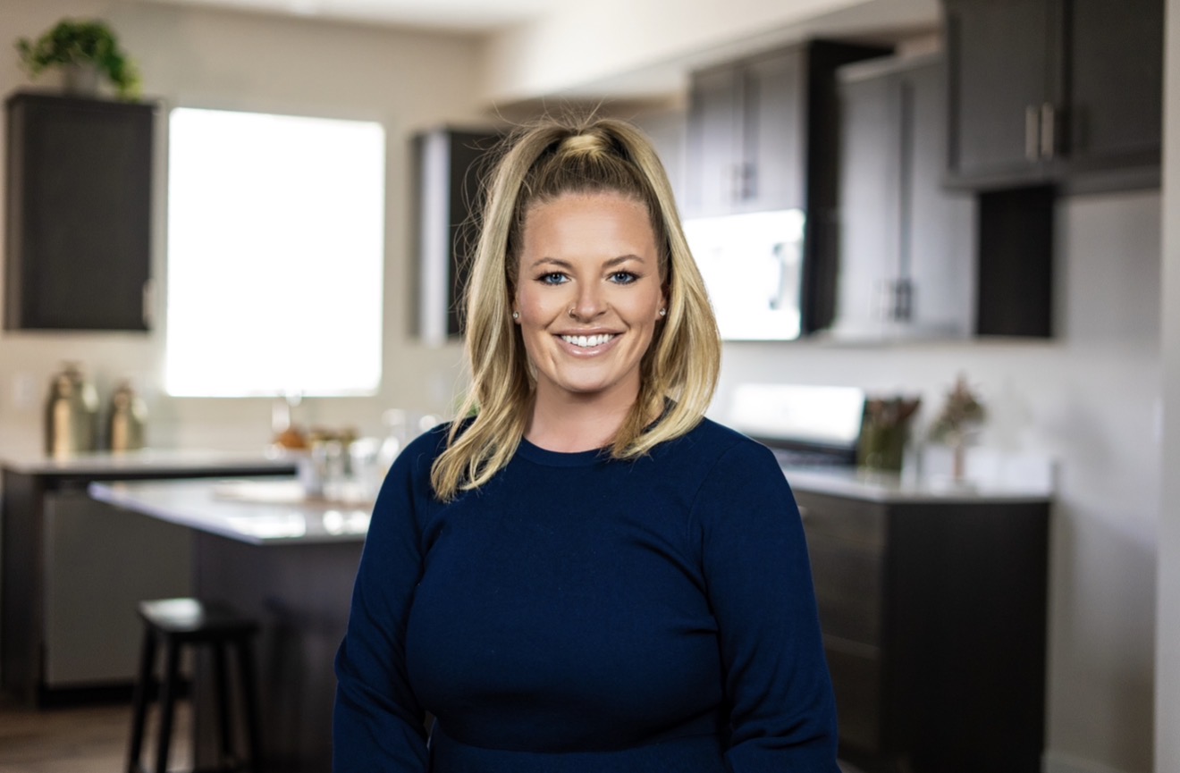 Smiling woman with shoulder-length blonde hair in a blue top standing in a modern kitchen.