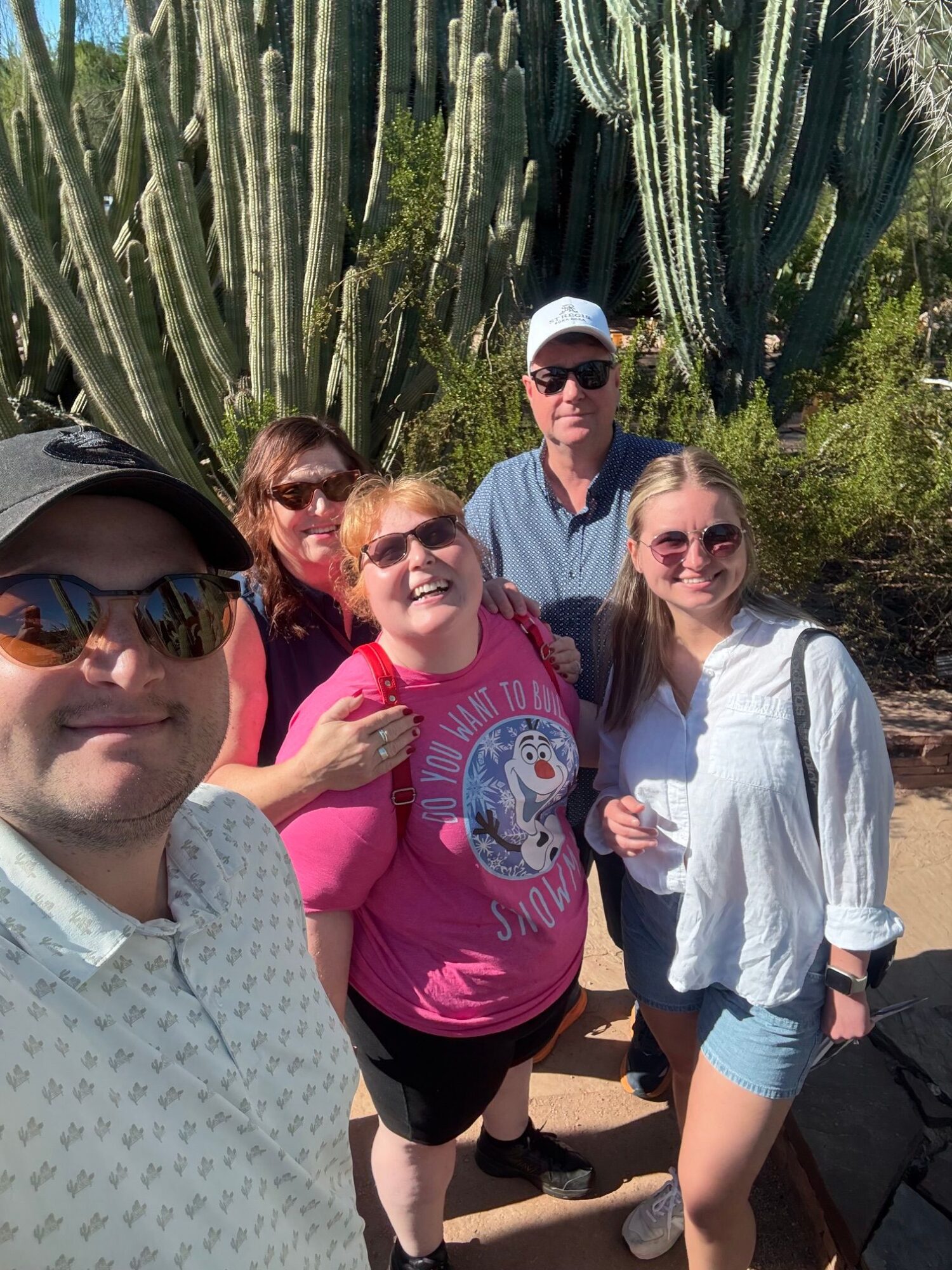 Four people standing outdoors near tall cacti, smiling and posing for a photo, with greenery in the background.
