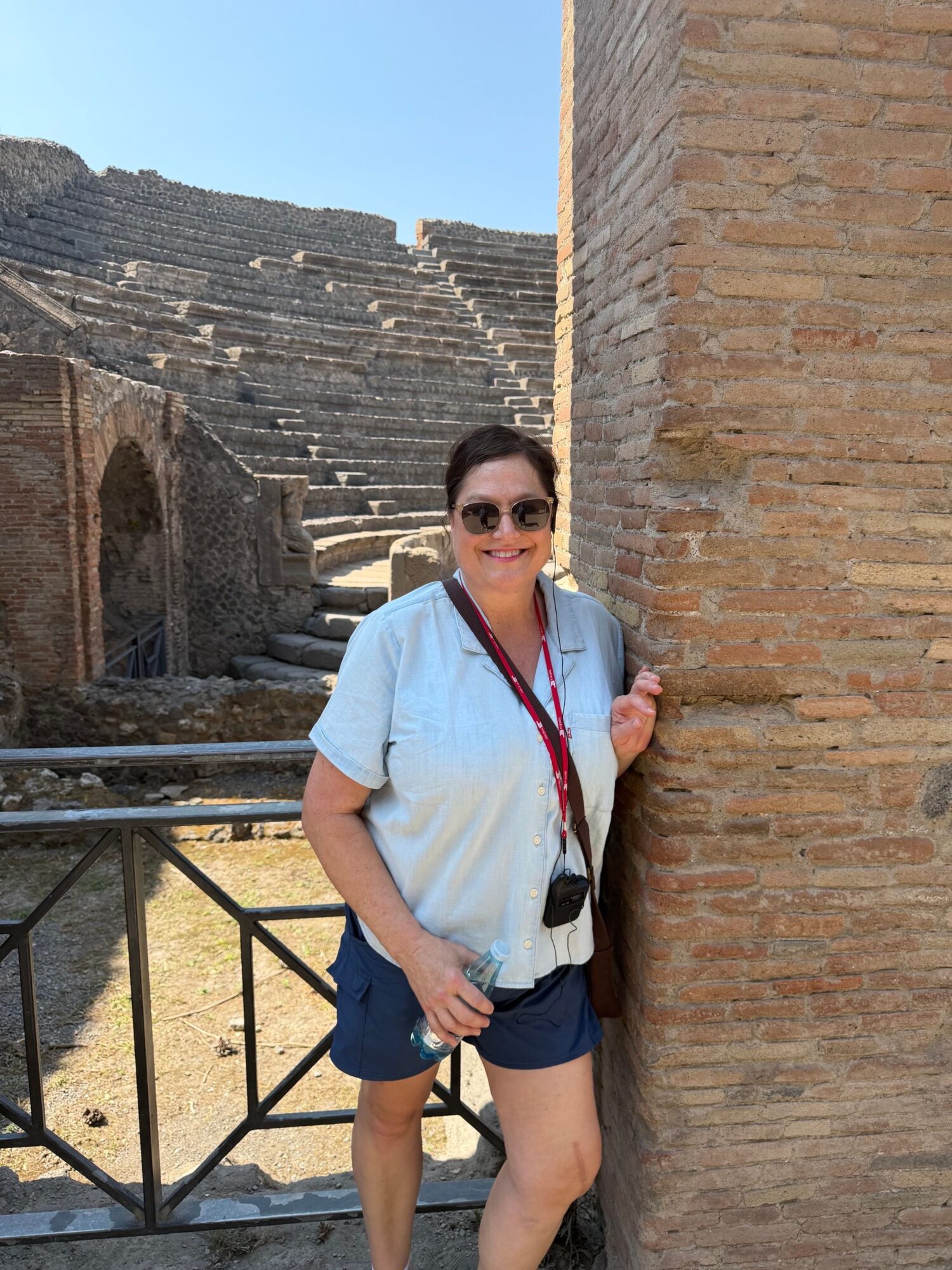 Woman standing outdoors near stone stairs, wearing sunglasses, a light blue shirt, and dark shorts, holding a bag strap.