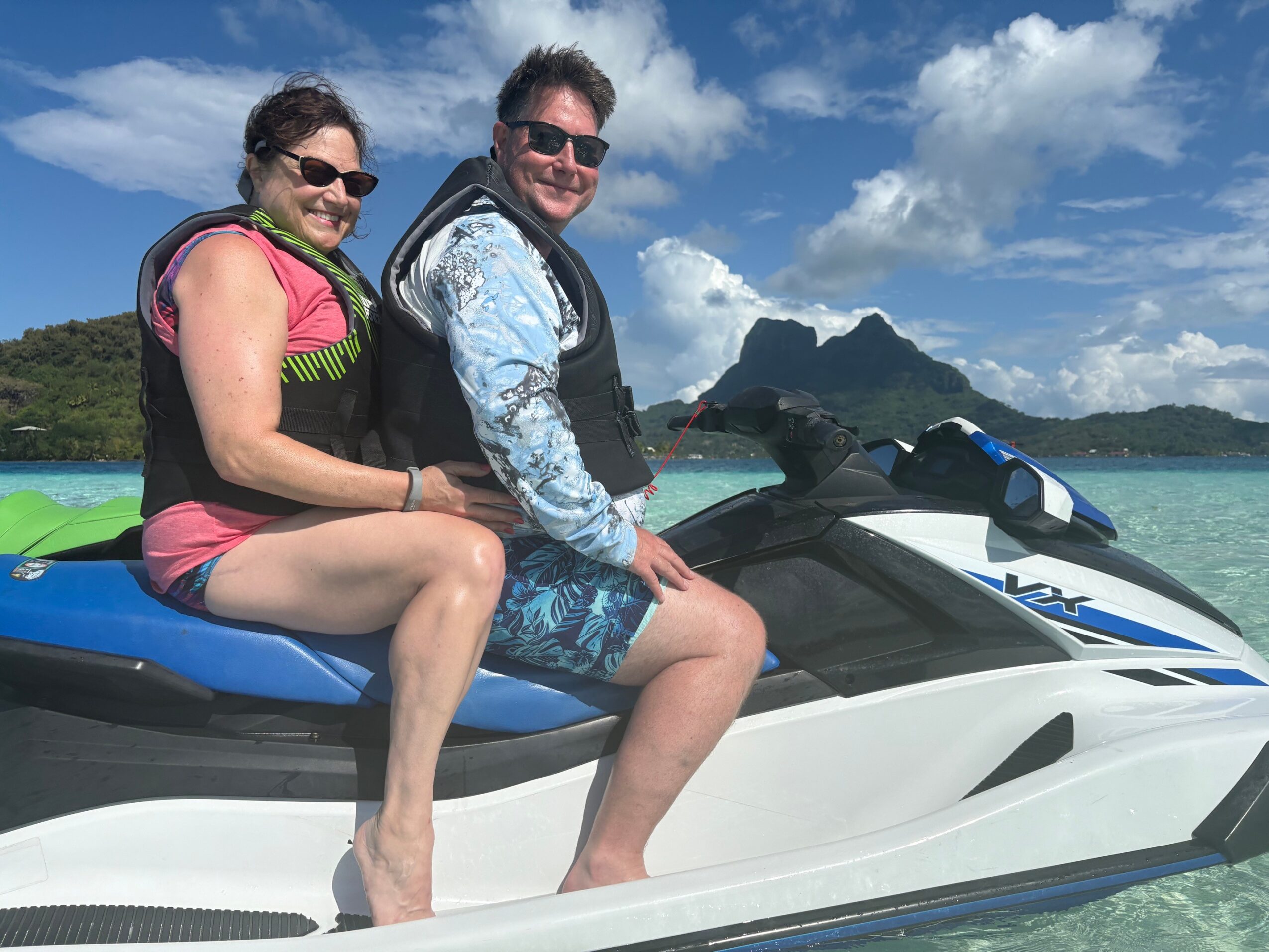 Two people sitting on a jet ski with a tropical island and mountain in the background, smiling at the camera.