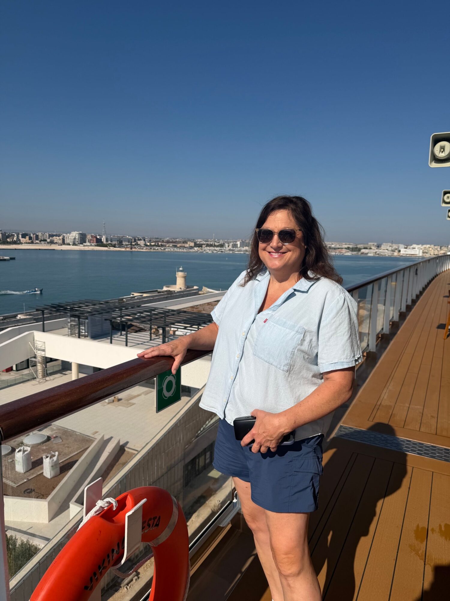 Woman standing on a boat deck with water and boats in the background, smiling, holding a phone, wearing sunglasses and casual clothes.