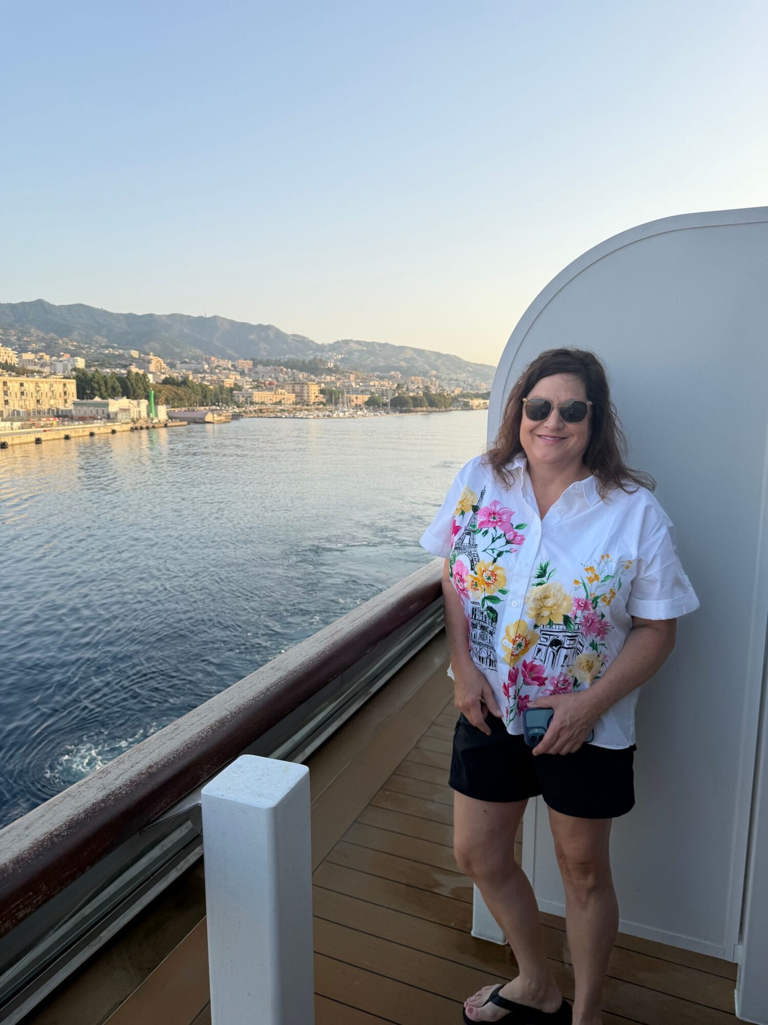 Woman standing on a boat deck with water and cityscape in the background, wearing a floral top and black shorts.