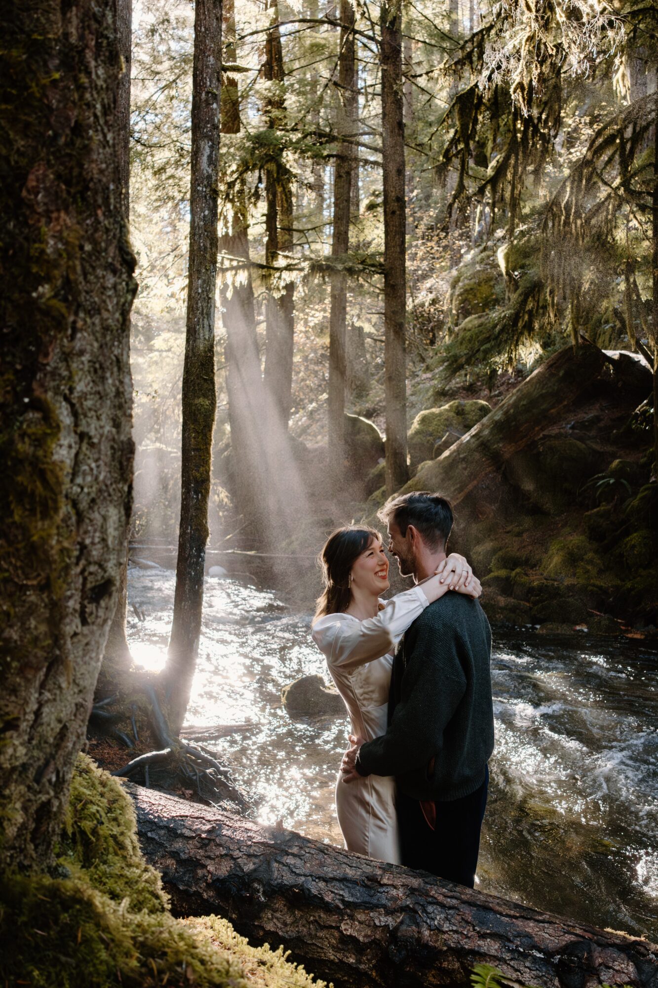 A couple stands in a forest near a stream with sunlight streaming through trees, a waterfall in the background.