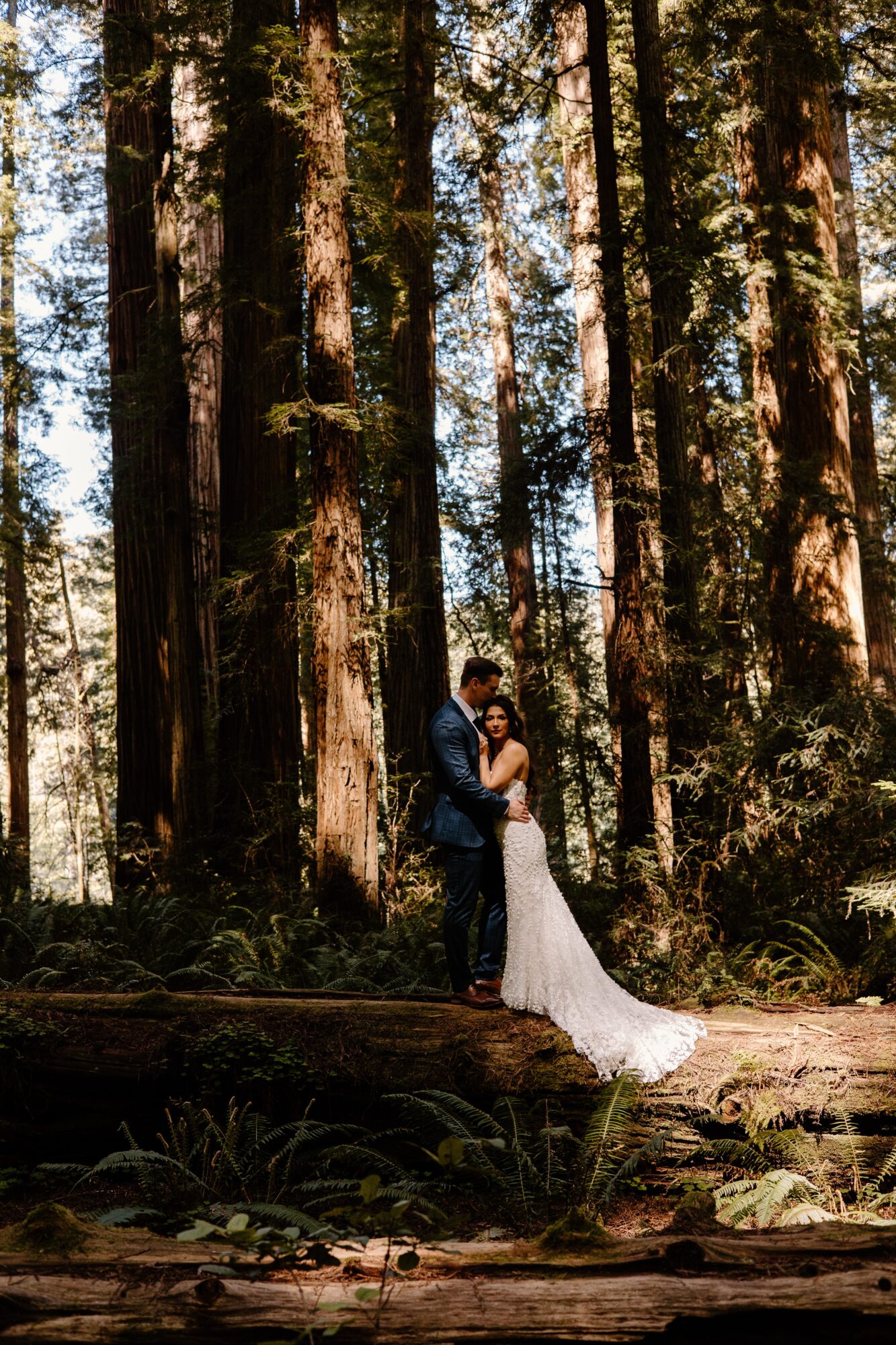 A couple stands close together in a forest with tall trees and sunlight filtering through the leaves.