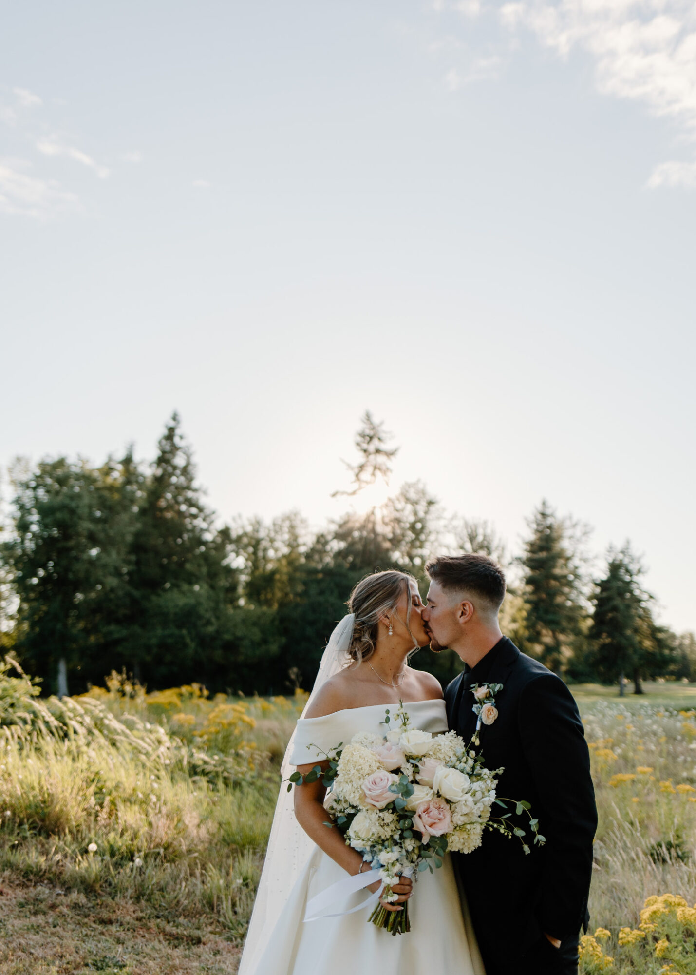 A bride and groom stand close together outdoors, with trees and a bright sky in the background, sharing a moment.
