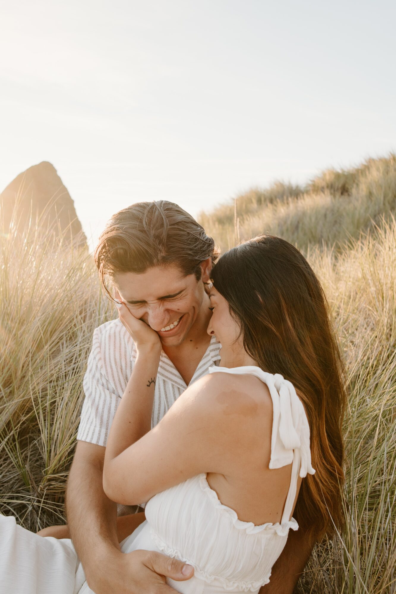 Two women sitting in tall grass, smiling and embracing outdoors on a sunny day.