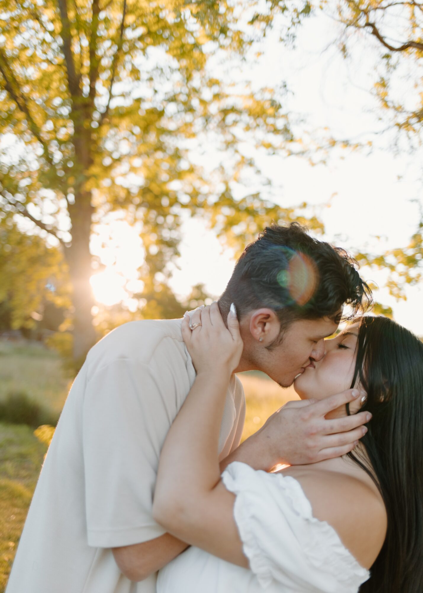 A couple kissing outdoors with trees and sunlight in the background.