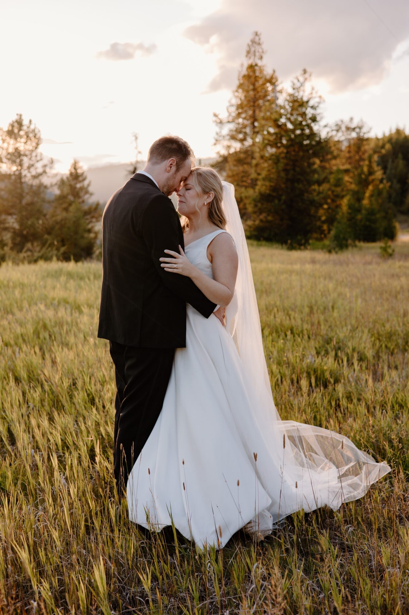 A bride and groom stand close in a grassy field, embracing each other during sunset, with trees in the background.