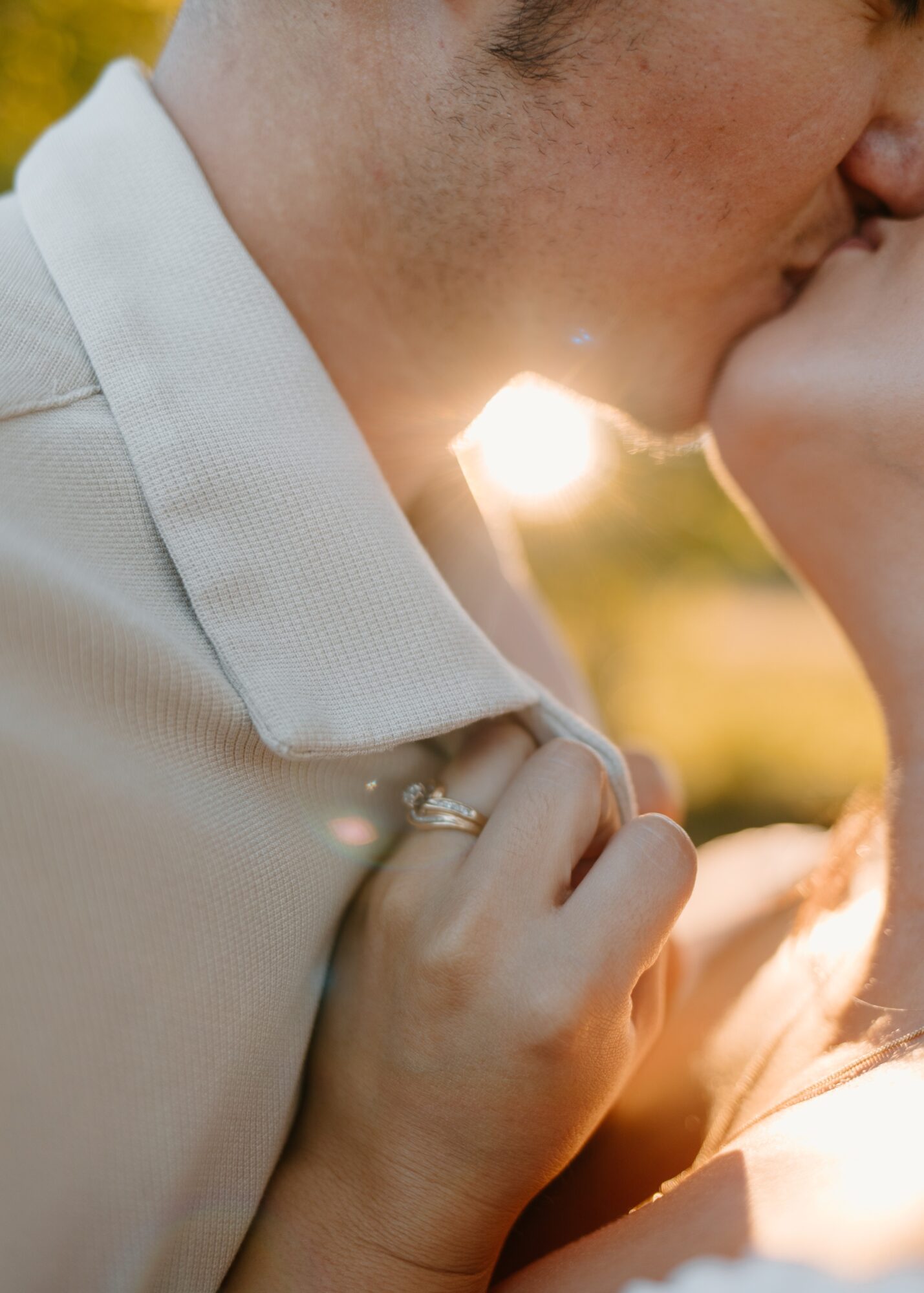 Close-up of a couple kissing outdoors, sunlight shining between them, woman holding man's collar, wedding ring visible.