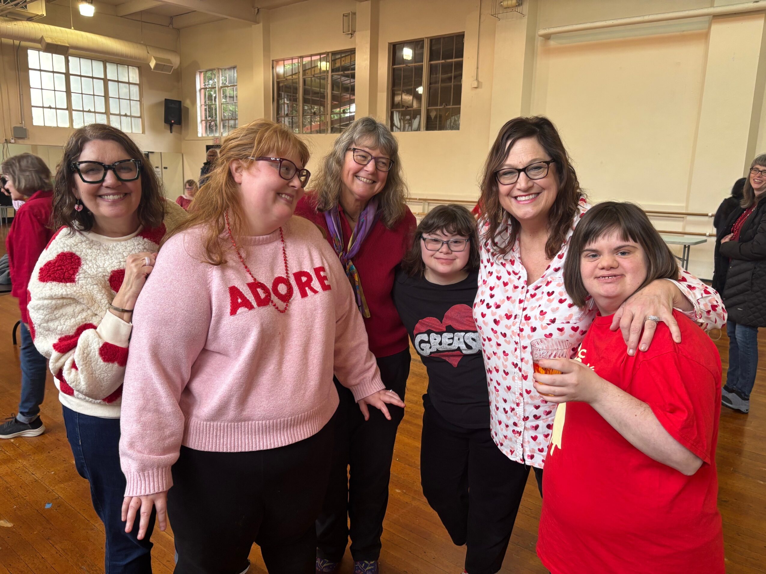 Group of six smiling women and girls standing indoors, some holding drinks, in a room with large windows and wooden floor.