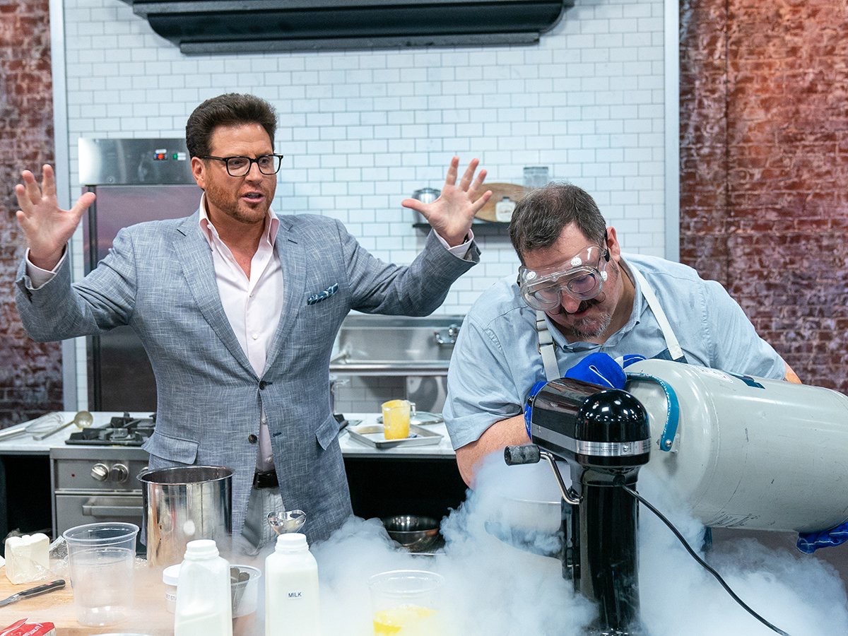 Two men in a kitchen, one speaking with hands raised, the other looking into a scientific instrument with smoke, glasses, and bottles around.