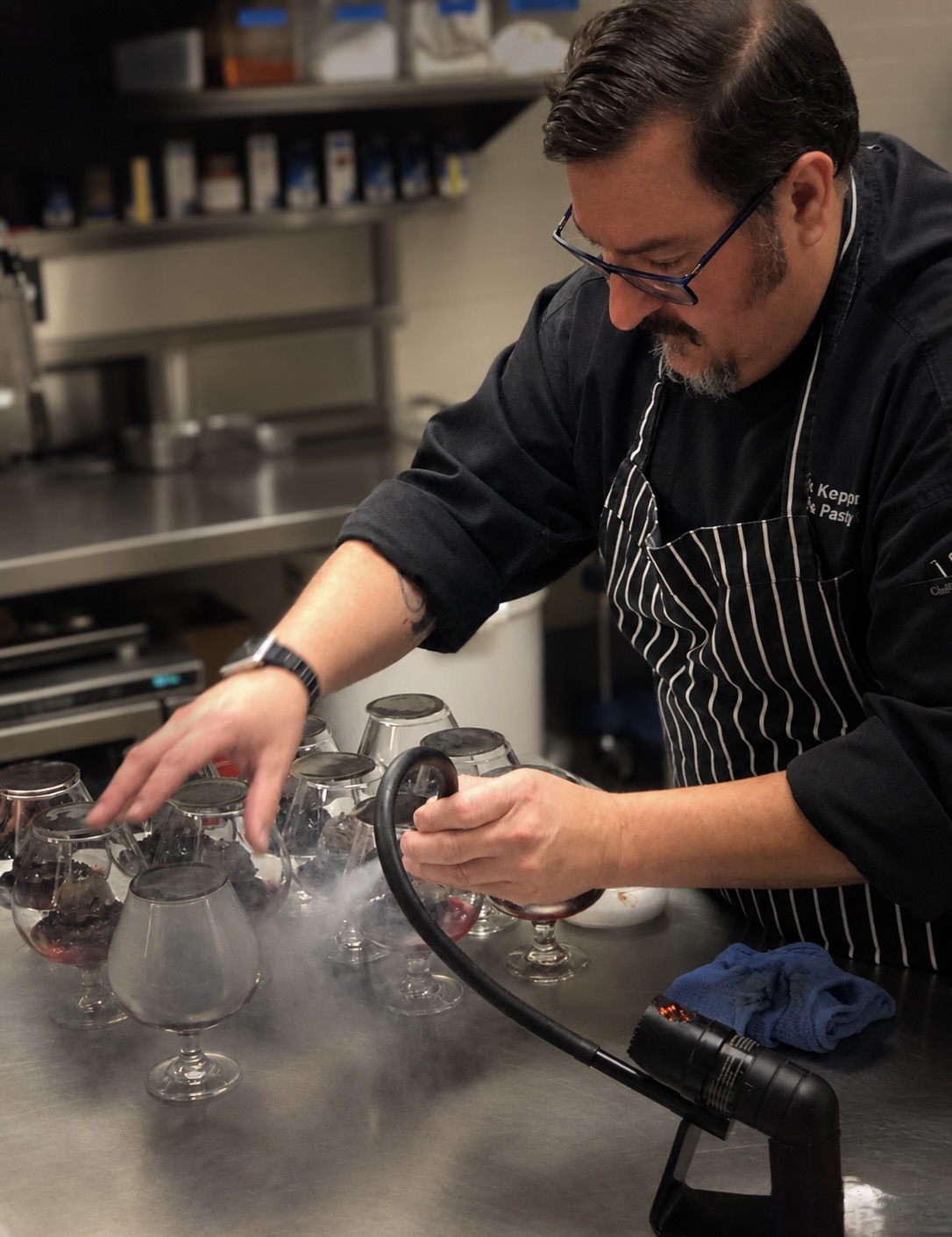 Chef in a kitchen, wearing glasses and a striped apron, handles glassware on a stainless steel counter.