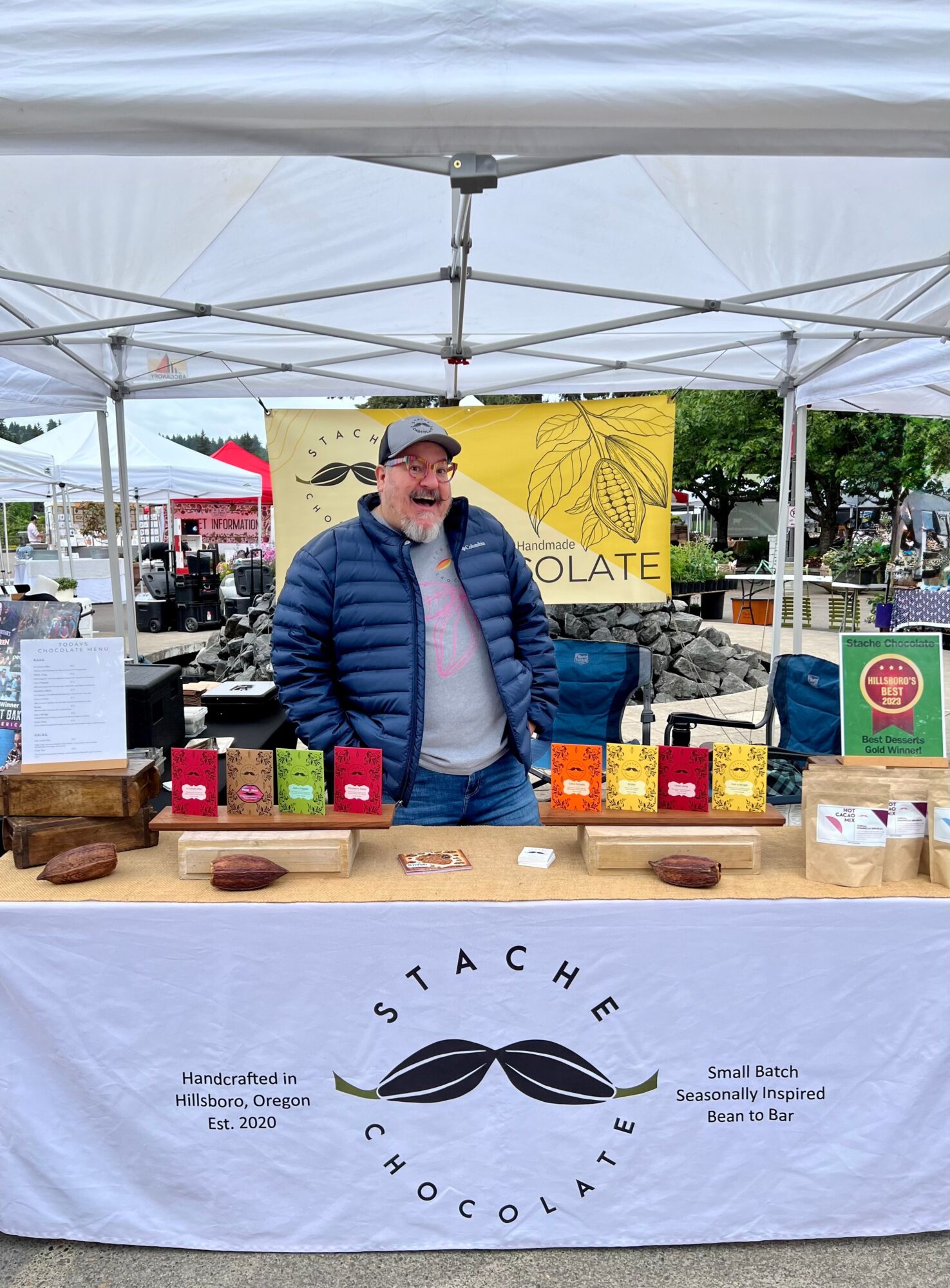 Man in blue jacket and cap standing behind a chocolate booth at an outdoor market, with a yellow sign and various chocolate products.