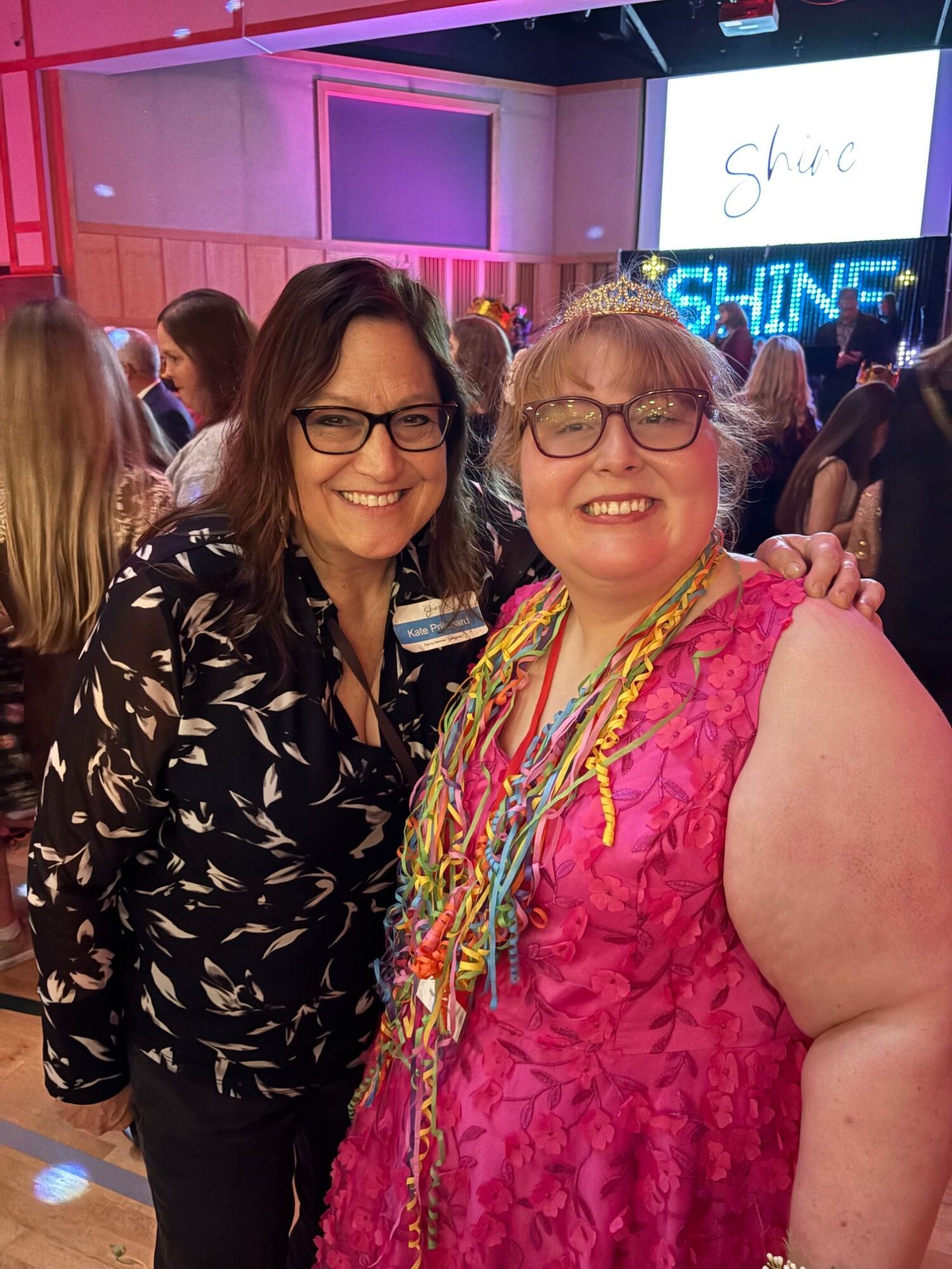 Two women wearing glasses and smiling, one in a pink dress and the other in a black patterned outfit, at an indoor event.
