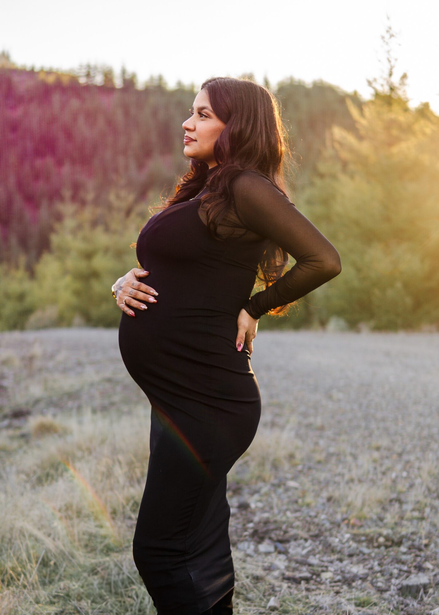 Pregnant woman in a black dress standing outdoors on a gravel path with trees and sky in the background.