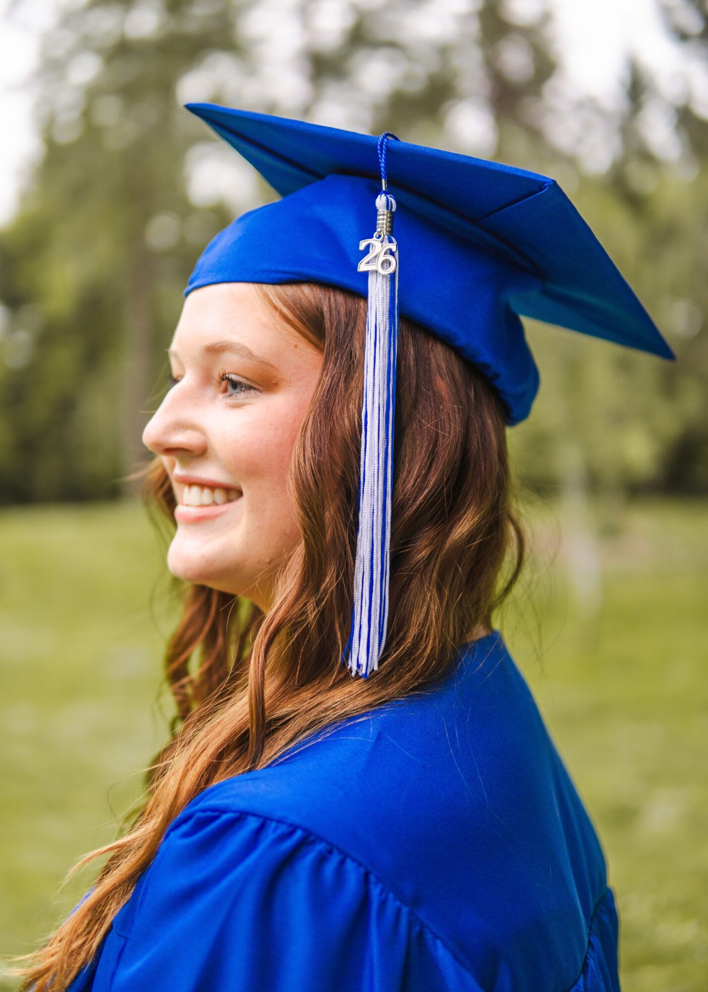 Young woman in blue graduation cap and gown outdoors, smiling, with trees in background.