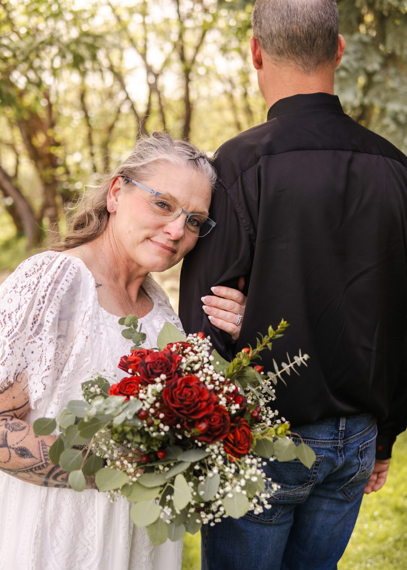 Young woman with glasses holding a bouquet of red and white flowers, standing outdoors with trees in background.