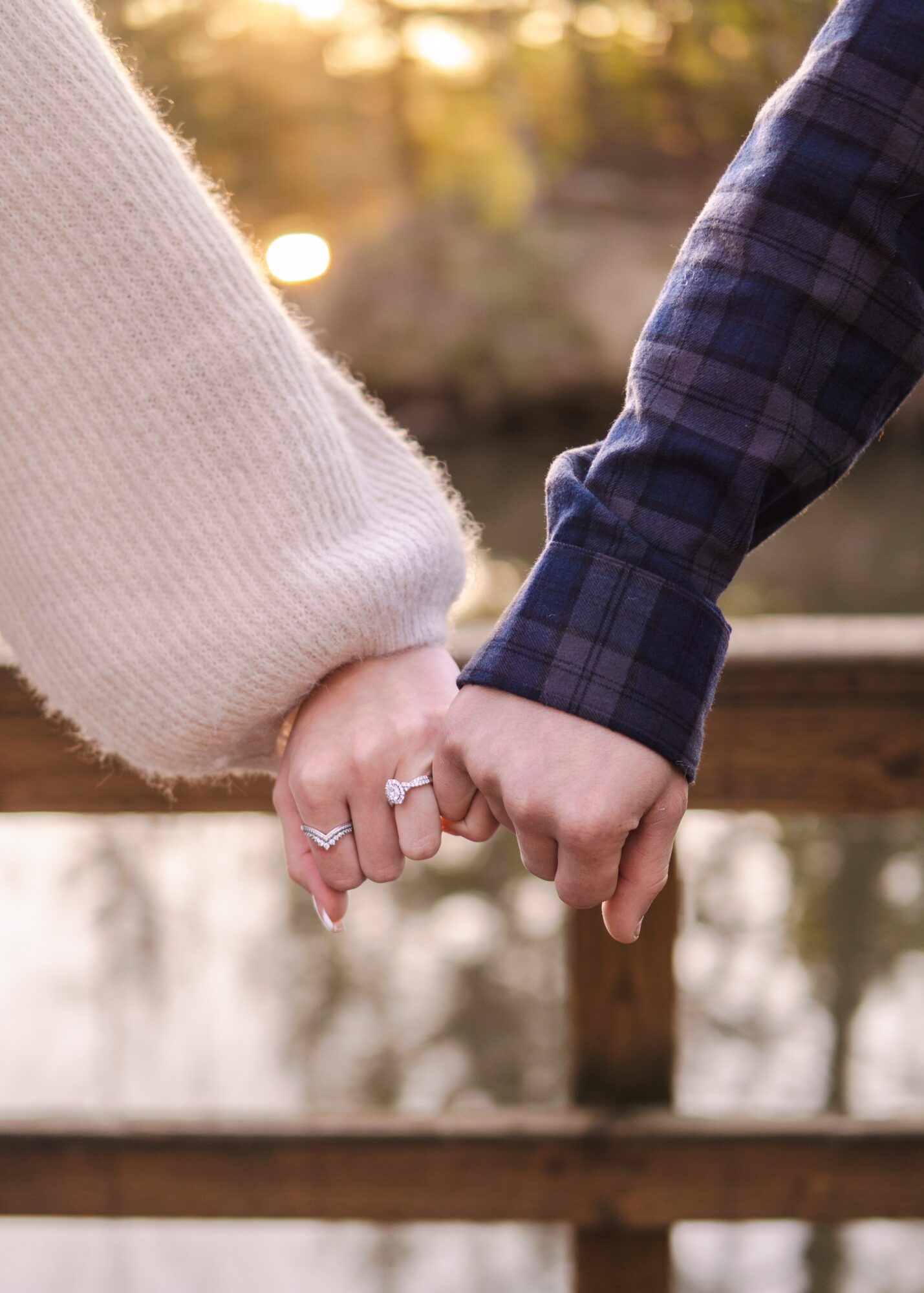 Two people hold hands, one wearing a white sweater and the other a blue plaid shirt, outdoors with blurred background.