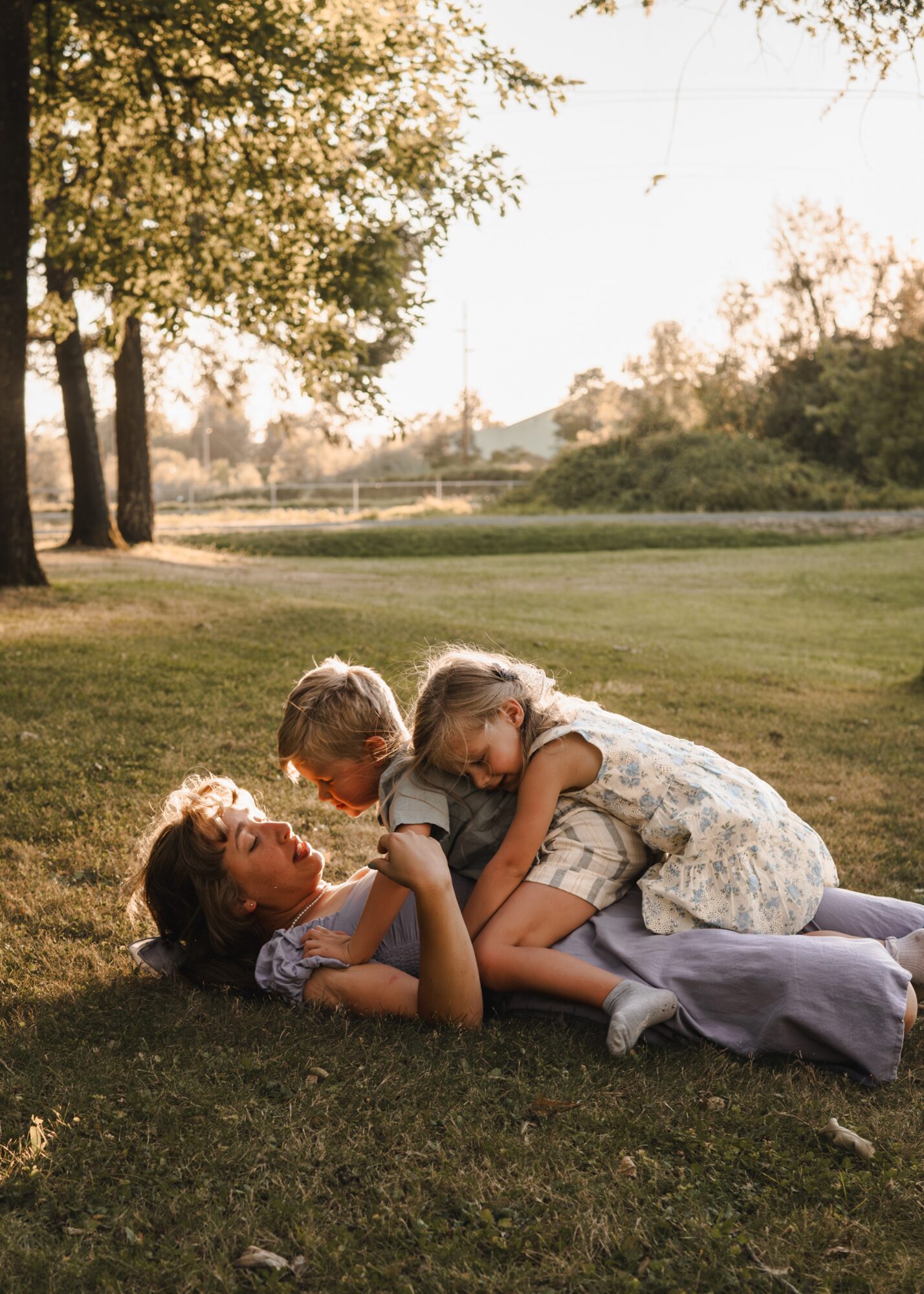 Two children playing on grass outdoors, one lying down and the other leaning over, with trees and a field in the background.
