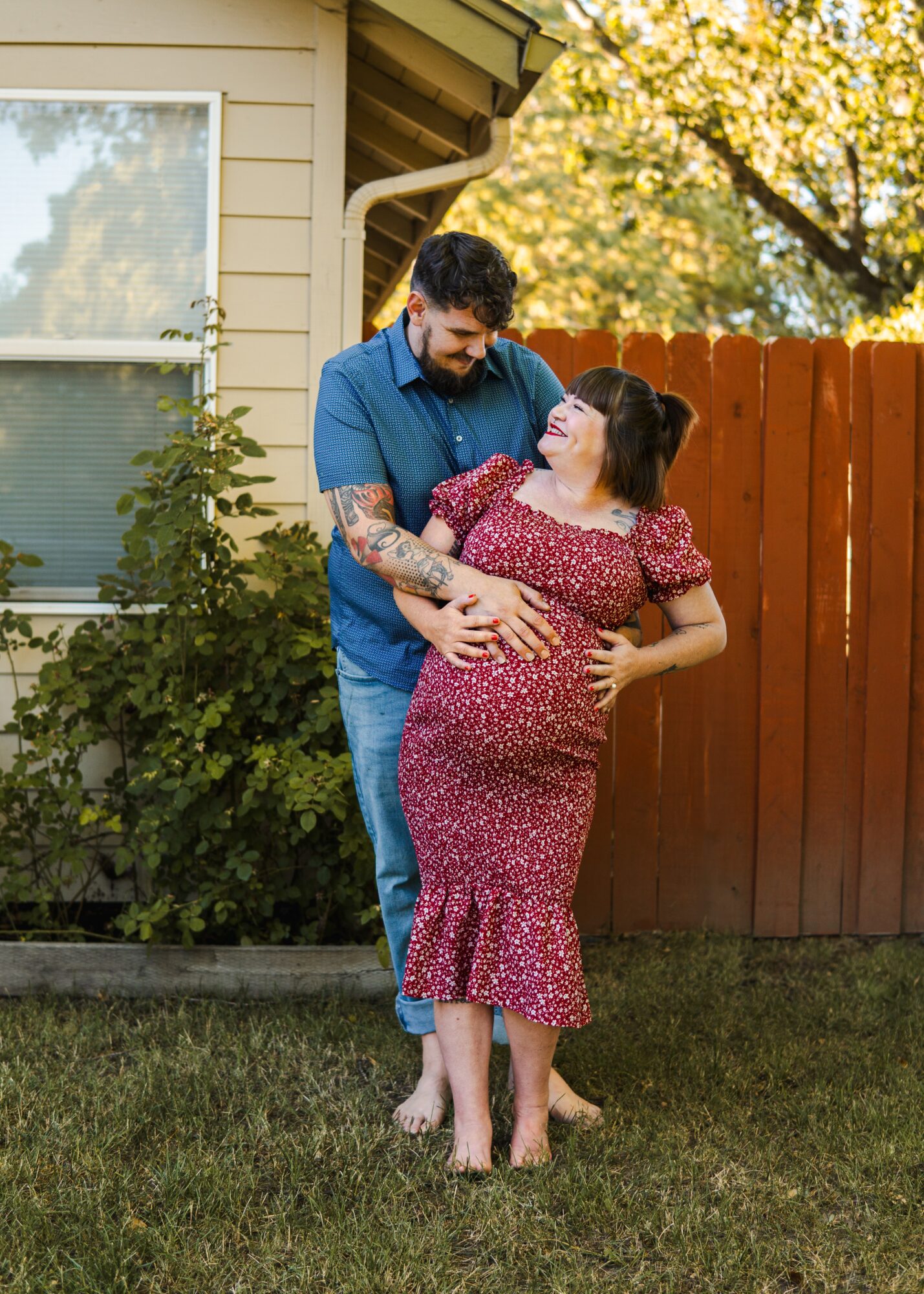 A couple stands outdoors near a house, embracing and smiling at each other, with trees and a fence in the background.