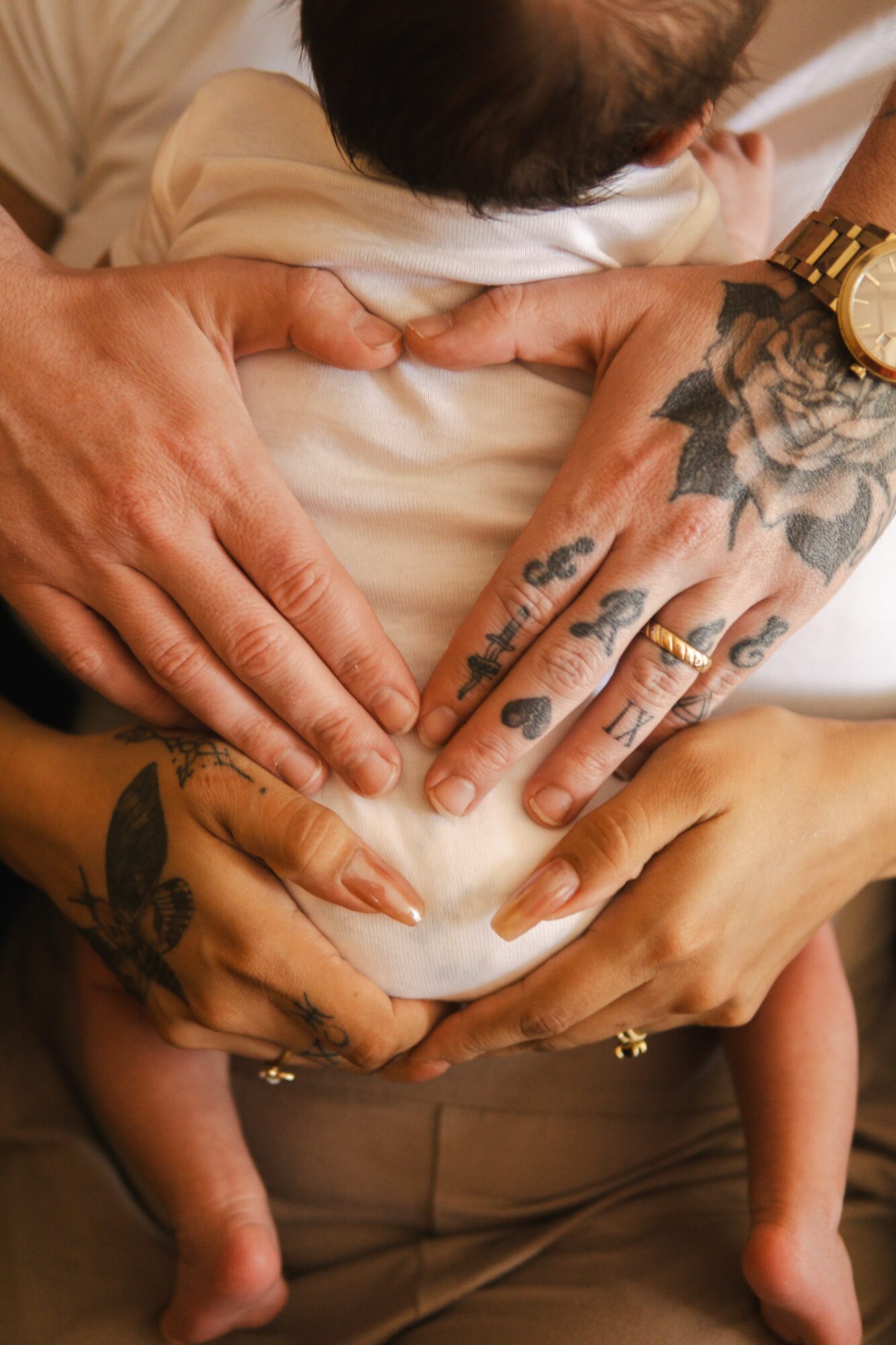 Two hands with tattoos hold a baby's torso, with the baby's legs hanging down. One hand has a gold watch.