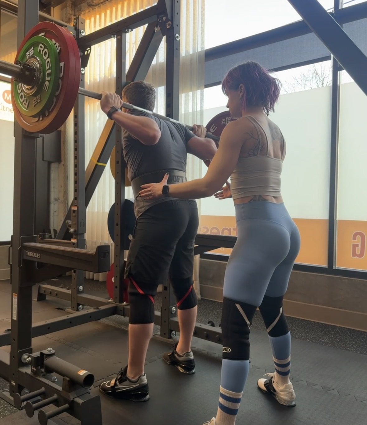 Two women in workout clothes lifting a barbell with weights in a gym, with large windows in the background.