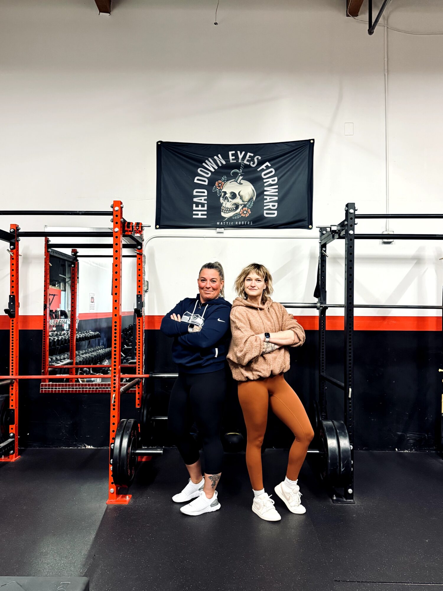 Two women stand in a gym, smiling, with a banner and gym equipment behind them.
