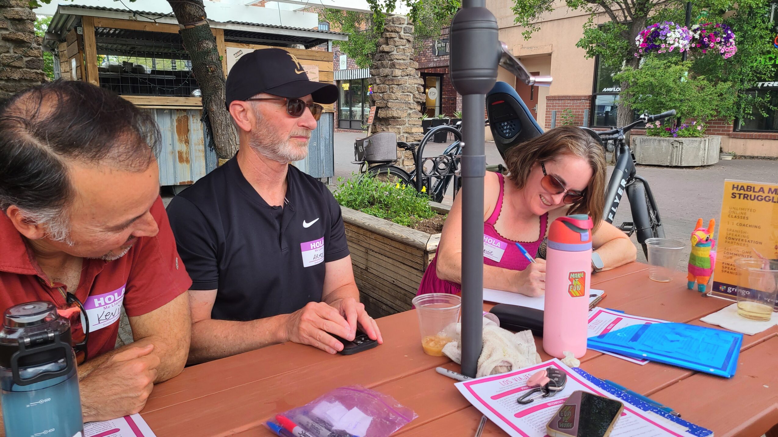 Three people sitting at a table outdoors, engaging with items and drinks, with trees and buildings in the background.