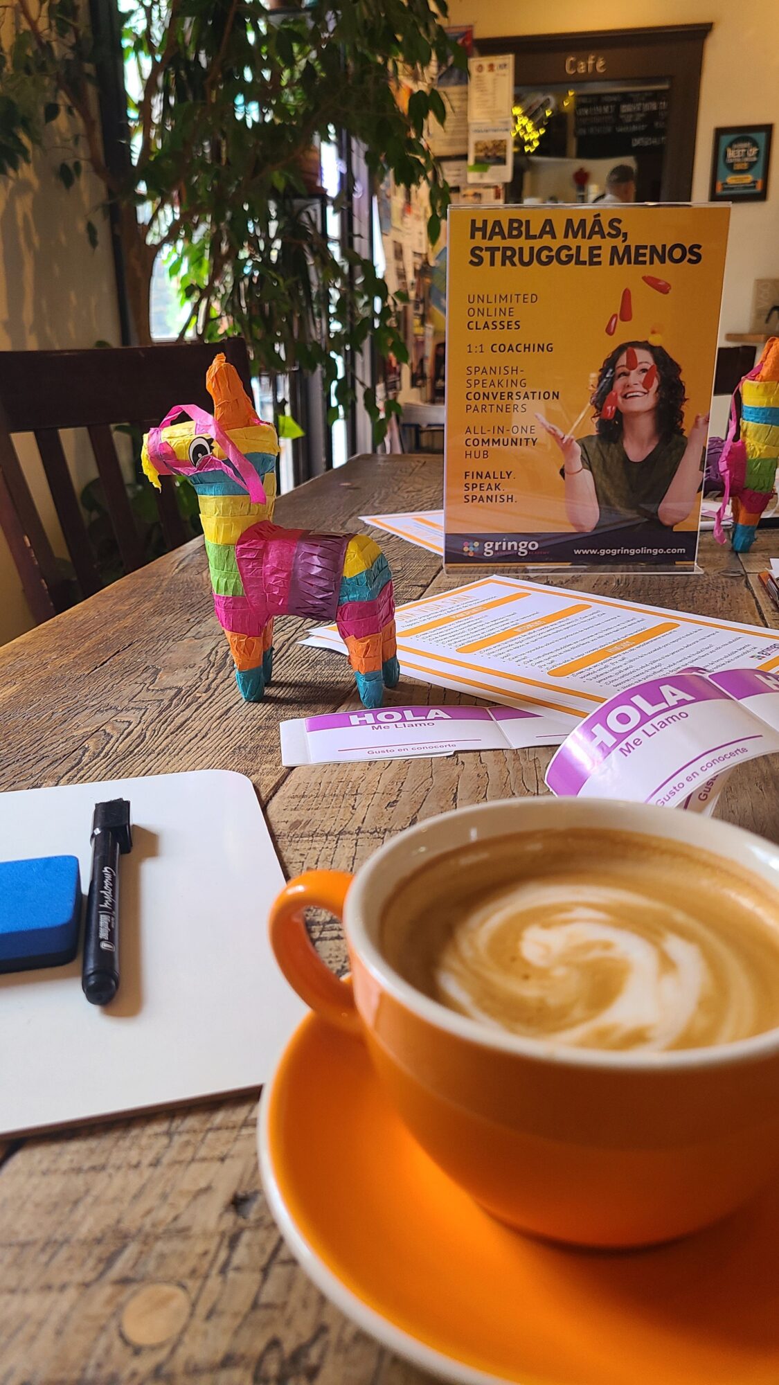 Coffee cup on table with promotional poster, colorful decorations, and a smartphone, outdoors setting with trees.