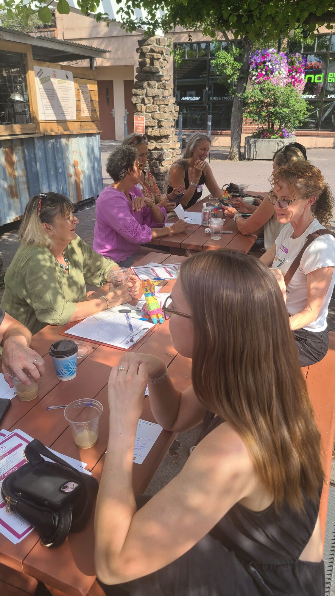 People sitting at a table outdoors, engaging in conversation and eating, with flowers and buildings in the background.