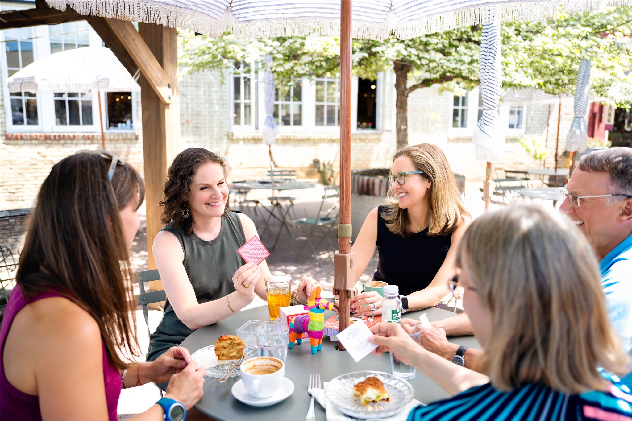 Group of five people sitting at an outdoor table, enjoying food and conversation, with trees and buildings in the background.